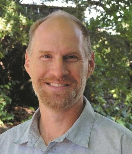 A smiling man with light skin, reddish-blond hair, and a goatee standing outdoors with green foliage in the background.