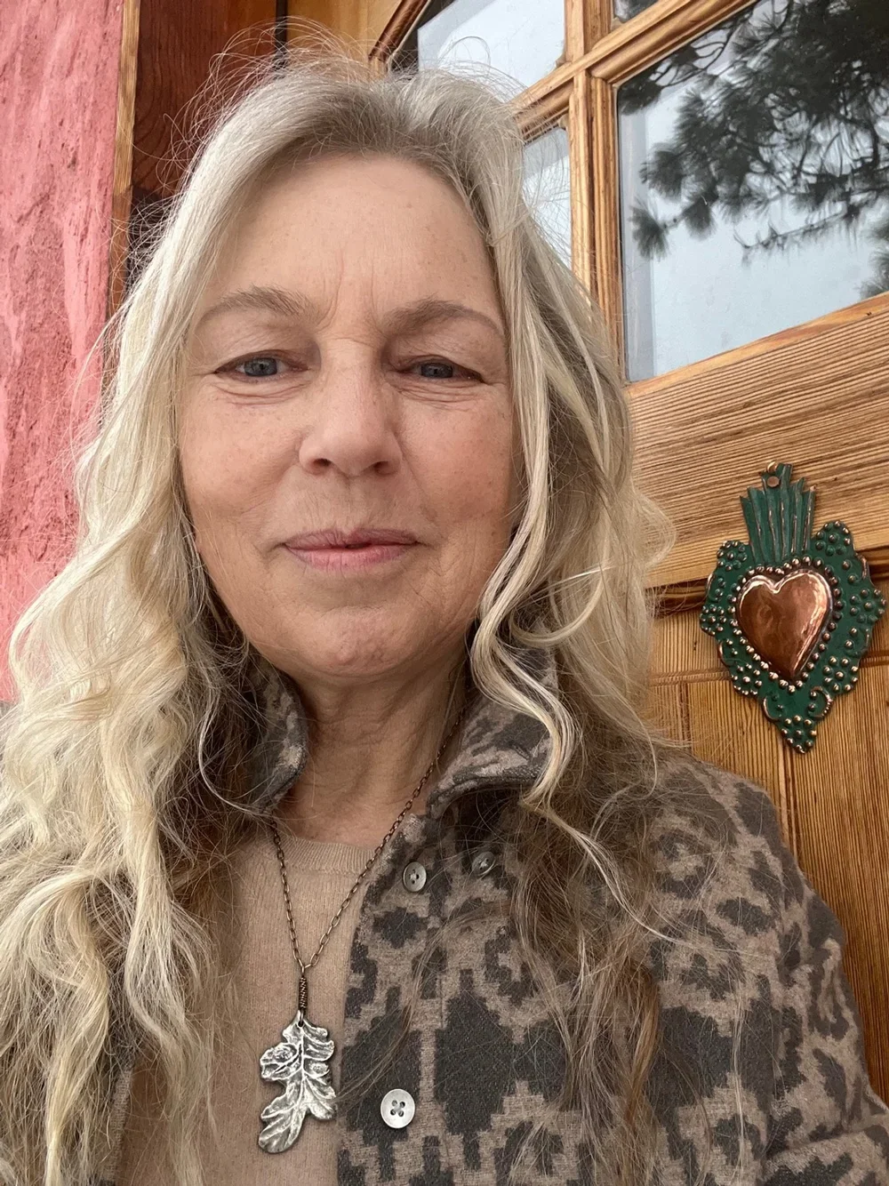A woman with long curly gray-blonde hair is smiling slightly at the camera. She is wearing a patterned jacket with a leaf-shaped pendant necklace. Behind her, there is a wooden window with a reflection of trees and a decorative metal heart on the wooden wall.