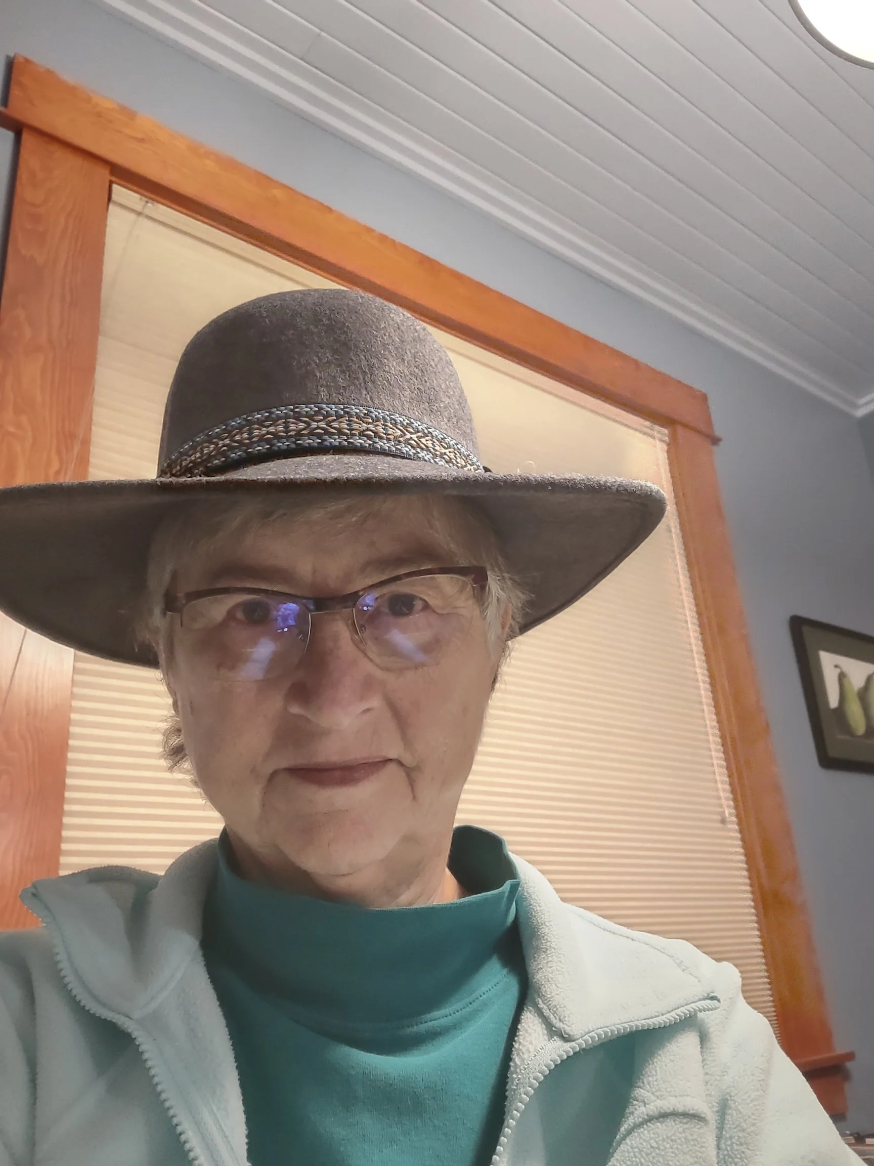 Close-up selfie of an older woman wearing glasses, a wide-brimmed hat with a decorative band, a teal shirt, and a light-colored jacket, standing in a room with wooden framing around a window and light blue walls.