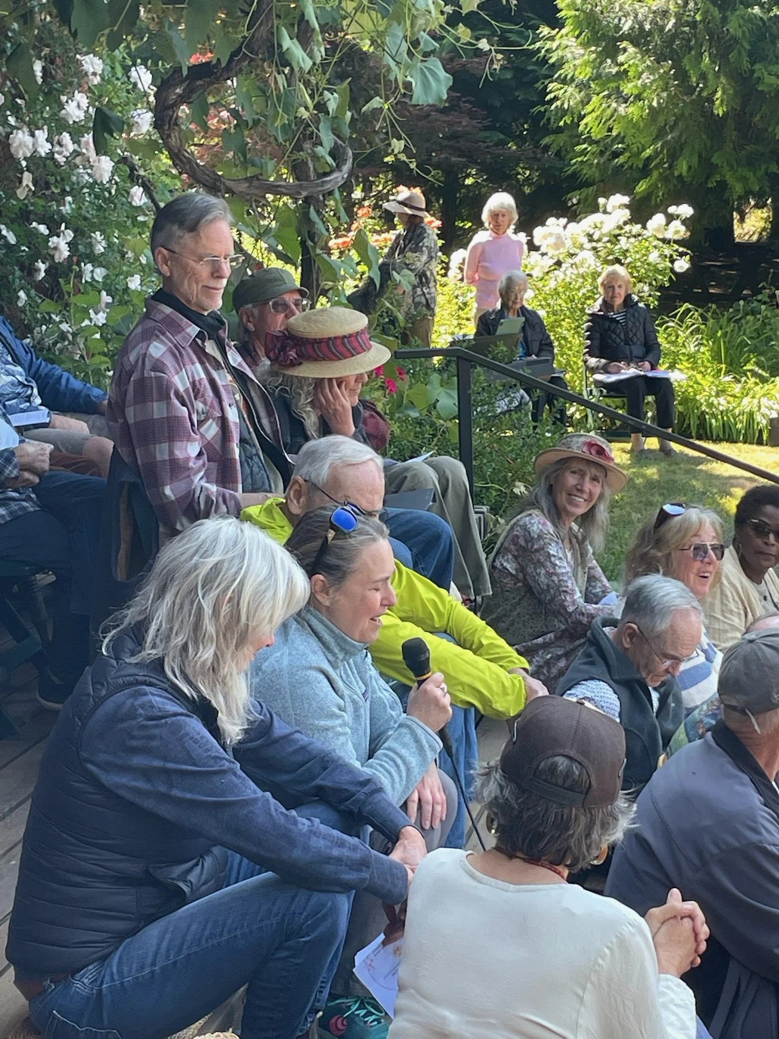 A group of people gathered outdoors, sitting and standing in meditation or prayer. The setting is lush with greenery and flowers, with some individuals wearing hats at our annual Flower Communion.