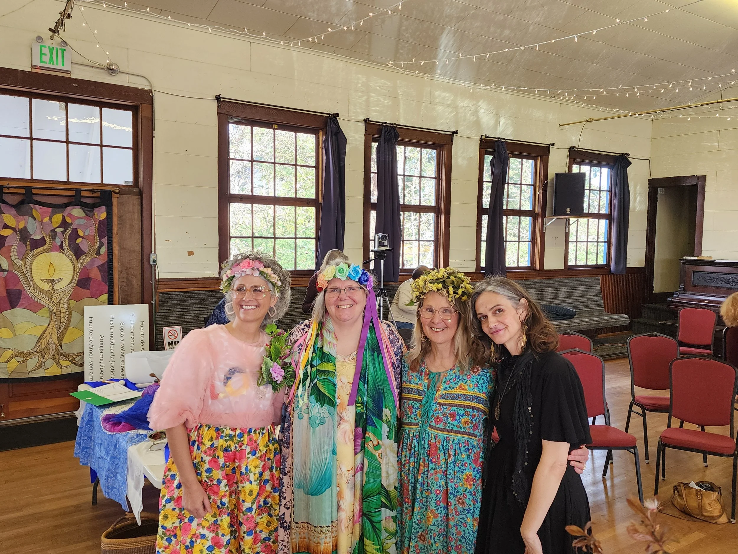 Four women are standing together and smiling at an indoor gathering, wearing colorful dresses and flower crowns. The background includes large windows, wooden window frames, string lights, and chairs.