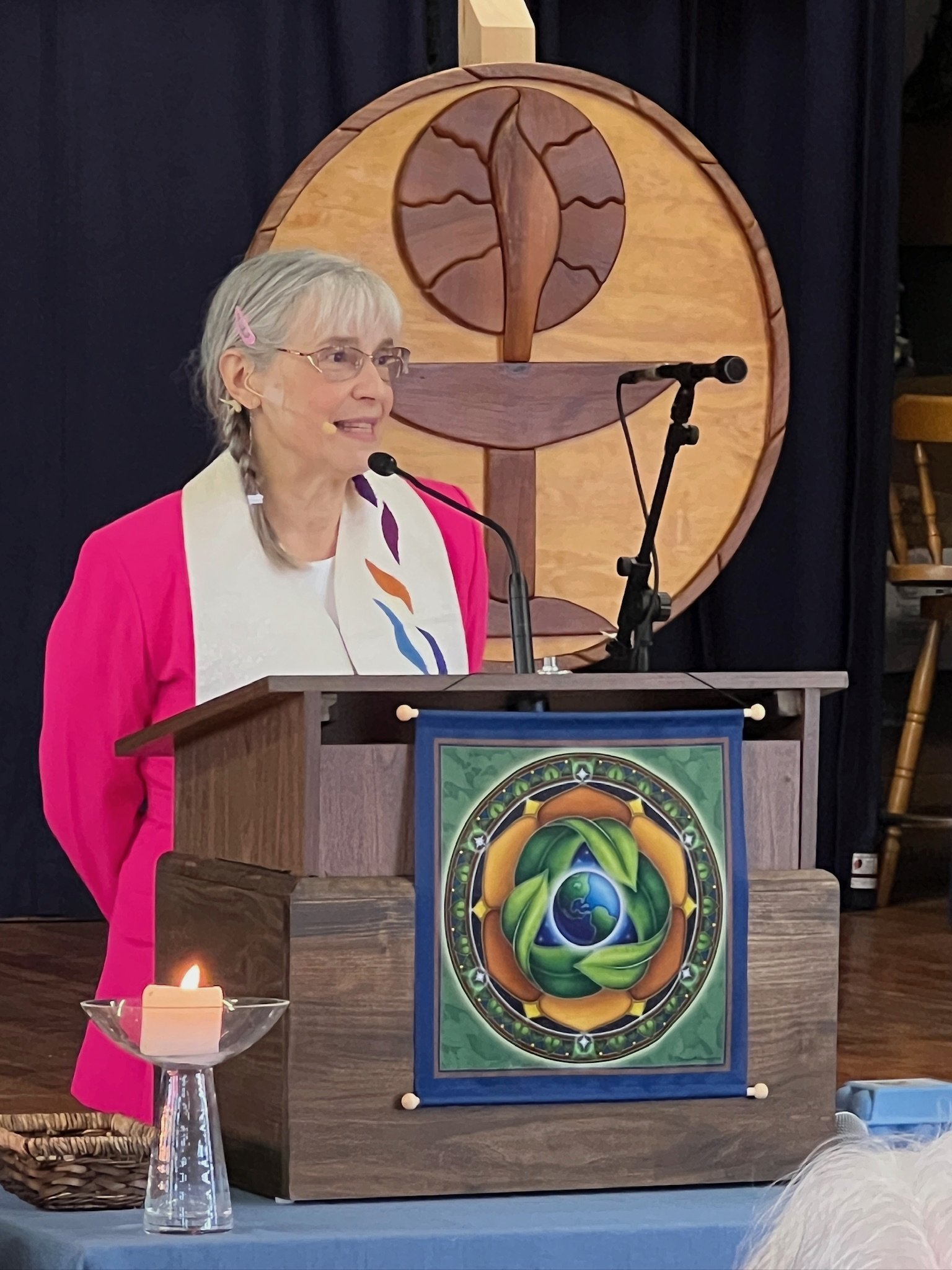 A woman with gray hair, glasses, and a pink blazer stands at a podium with a colorful circular emblem. Behind her is a wooden art piece resembling a tree with a circular background. There is a lit white candle in a glass holder on the table in front of her.