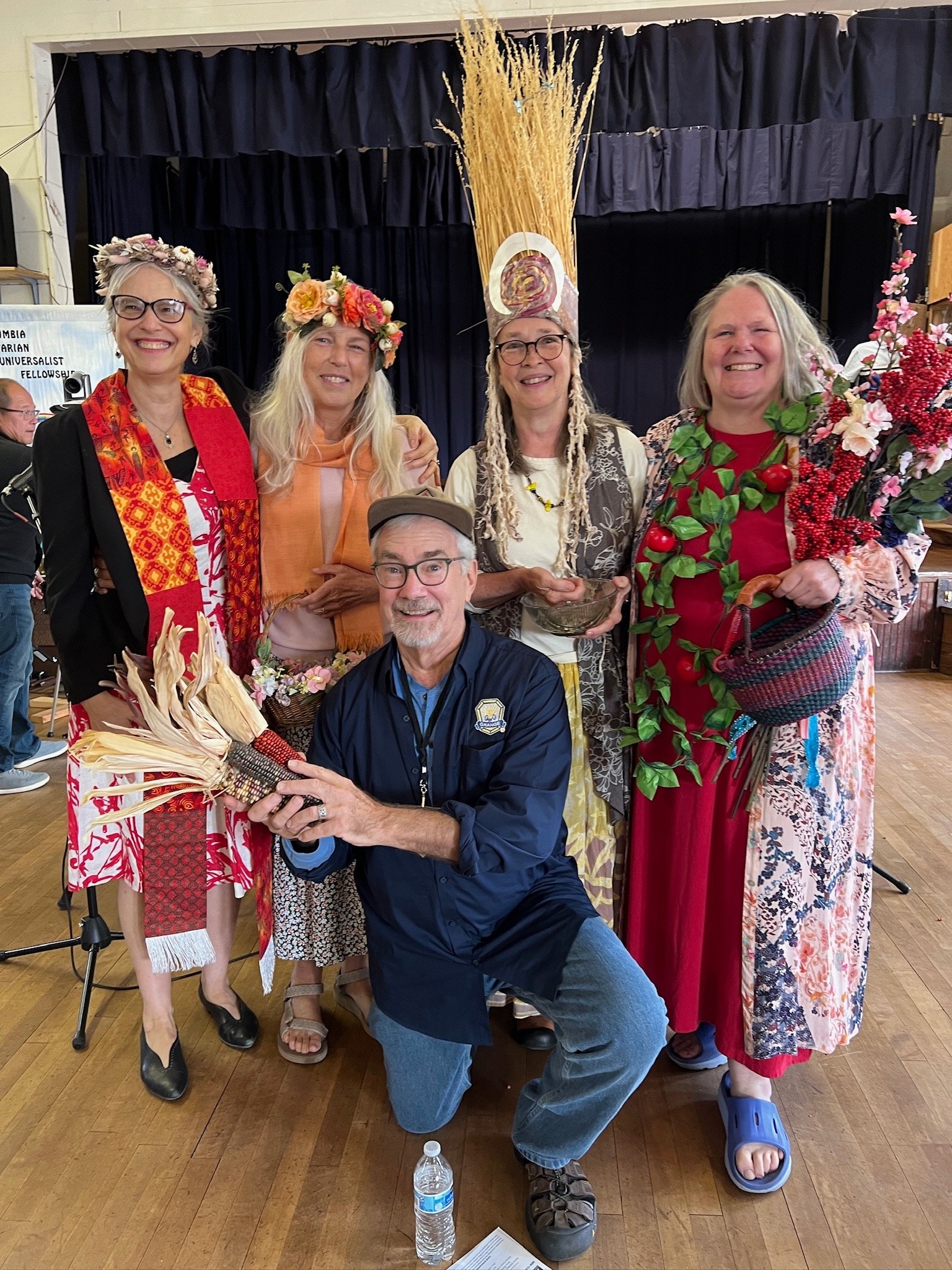 Group of five women and one man posing indoors, many wearing floral accessories and costumes, some holding flowers; one man kneeling in front with a water bottle nearby, smiling at the camera.