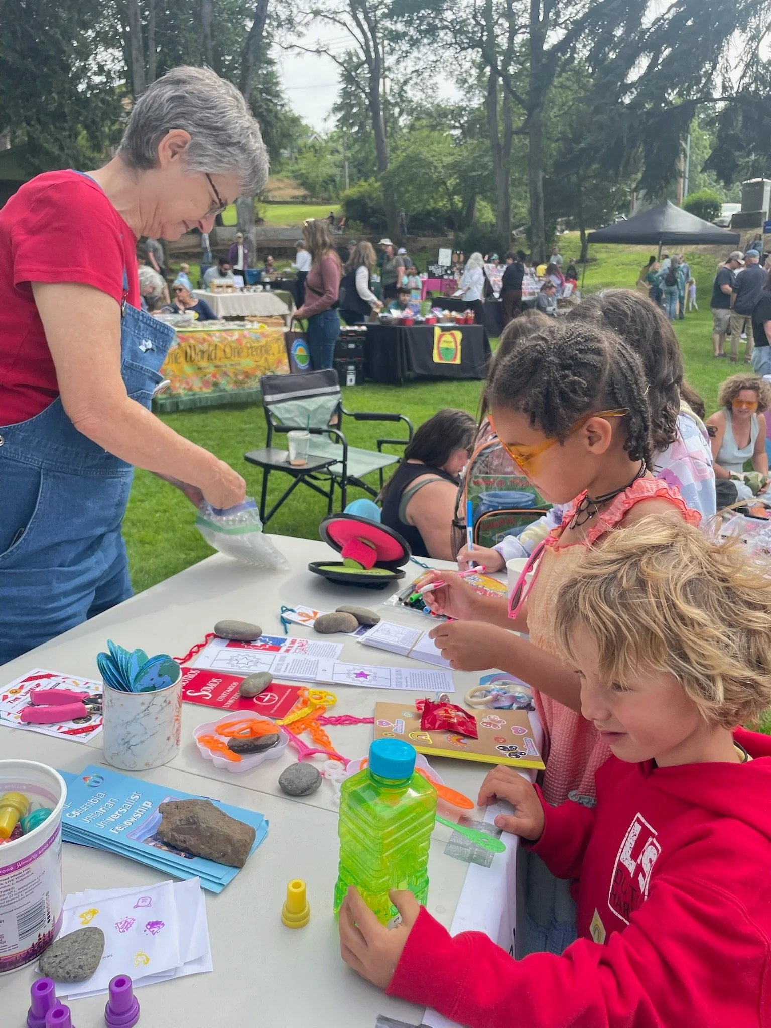 Children and adults engaging in arts and crafts activities at an outdoor event on a grassy area with trees. There are tables with supplies, rocks, and various craft materials. People are walking and browsing booths in the background.