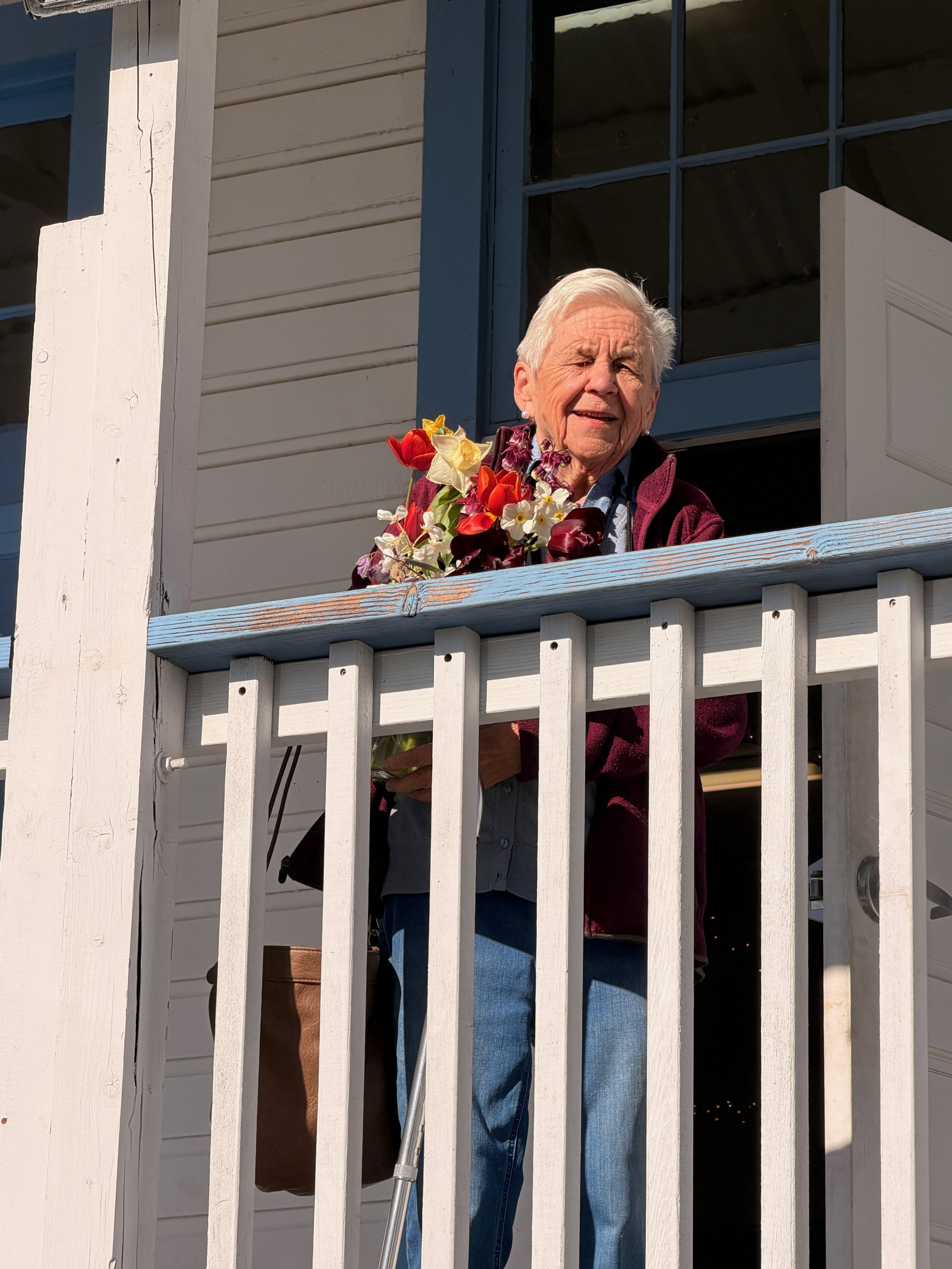 An elderly woman with white hair standing on a porch or balcony, holding a bouquet of colorful flowers, and smiling.