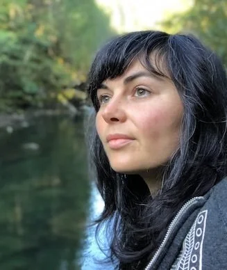 Young woman with black hair and light makeup looking to the right outdoors near a body of water with green trees in the background.