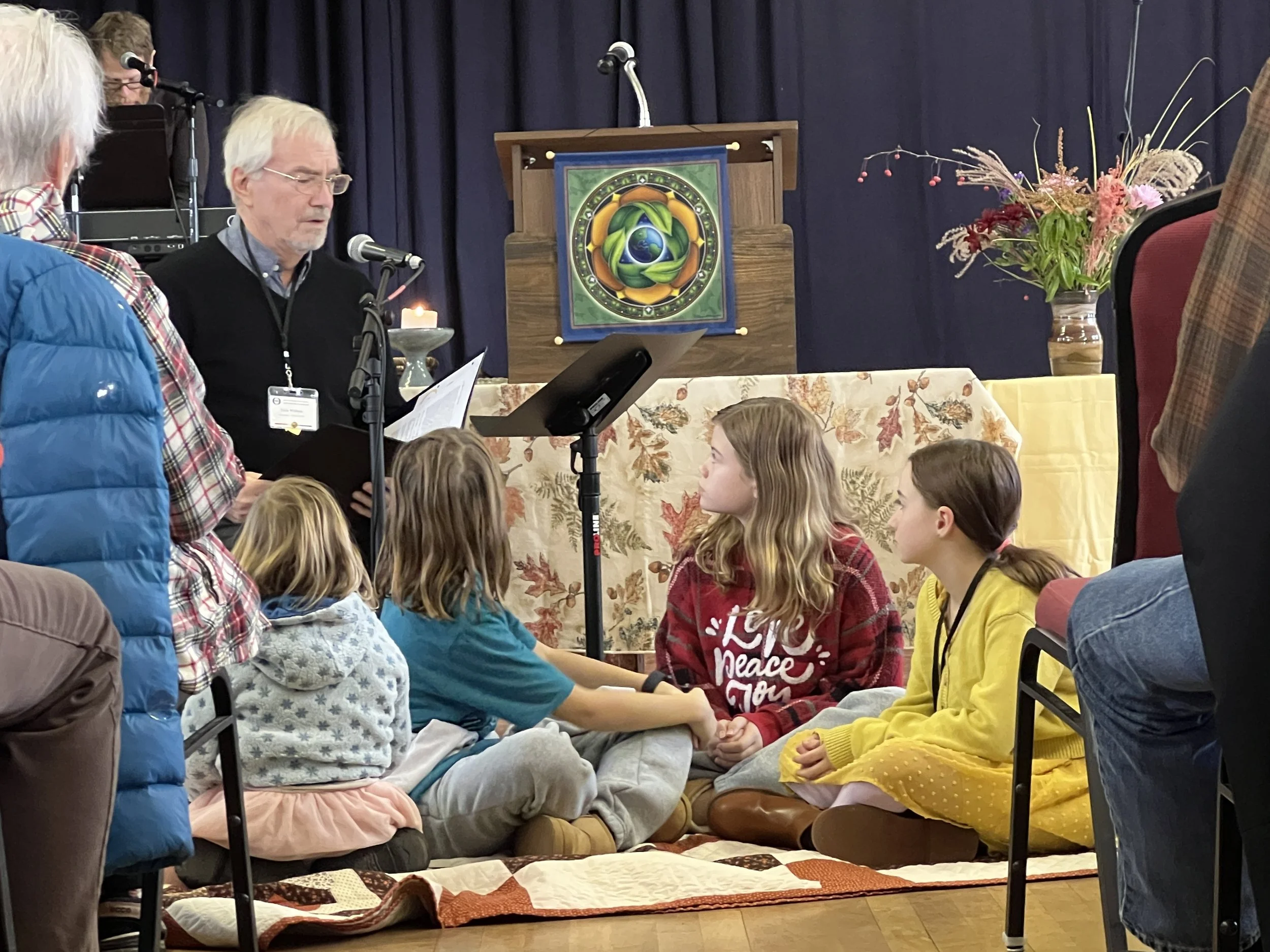 A man with gray hair, glasses, and a beard, wearing a black sweater, is speaking into a microphone, holding a paper, and standing at a table with children sitting cross-legged on the floor. The children are wearing colorful clothes; some have long hair, and one girl has a red sweater with white writing. In the background, there is a wooden stand with a colorful circular artwork, a flower arrangement, and a dark curtain.