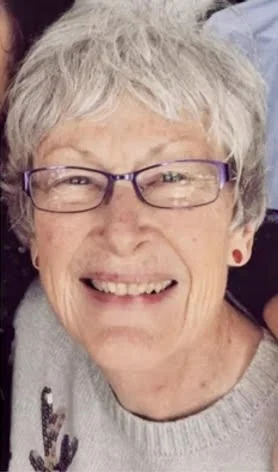 Close-up of a smiling older woman with short gray hair and purple glasses, wearing a light gray sweater.