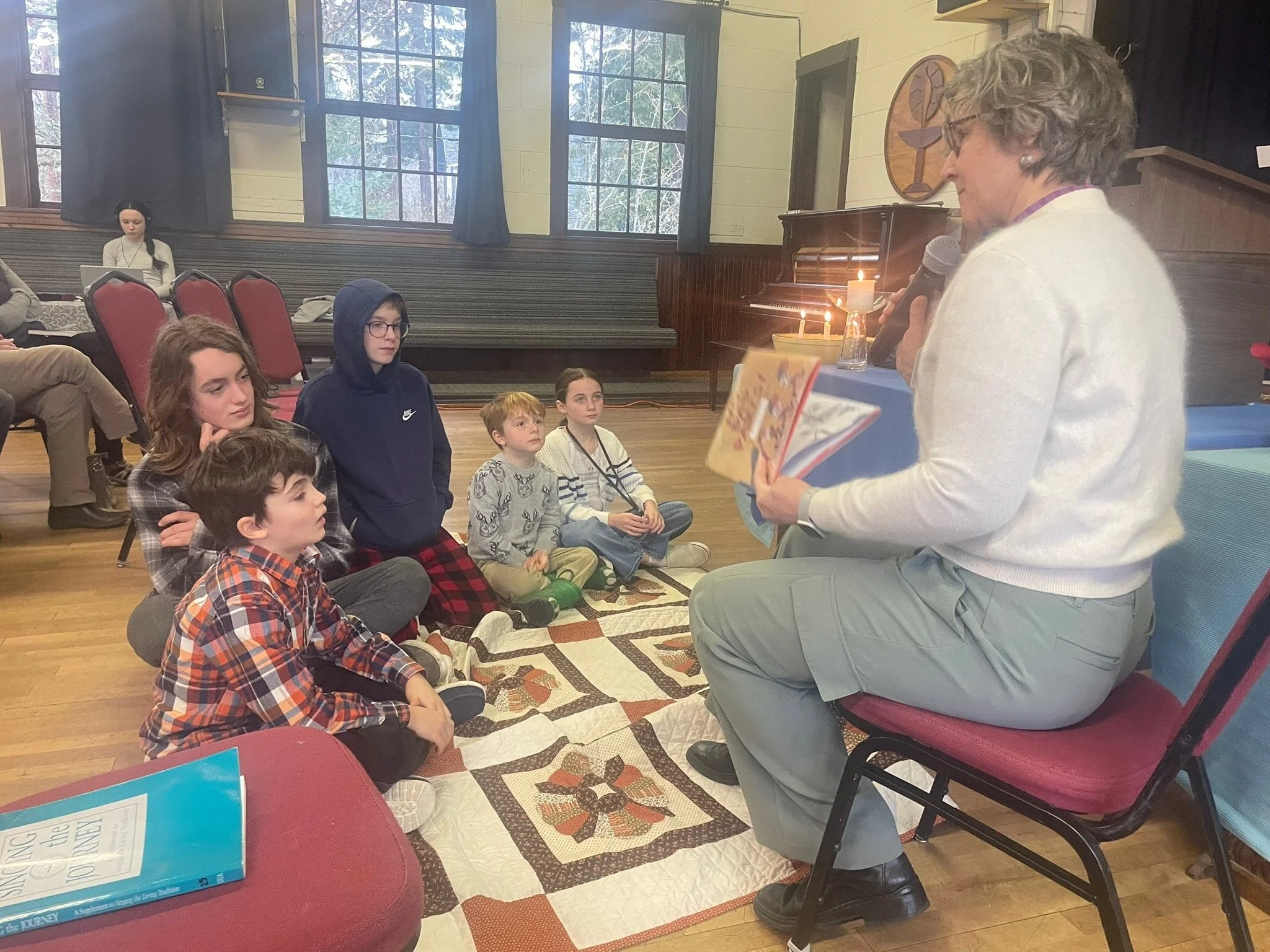 An older woman with short gray hair and glasses speaking into a microphone while holding a colorful booklet, with a group of children sitting on a patterned quilt on the floor, listening attentively inside a room with large windows and a piano in the background.