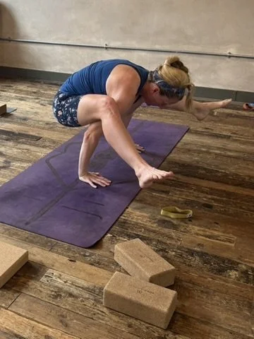 Lucy Tugwell practising firefly pose in a yoga studio, balancing on her hands and feet on a purple yoga mat, with yoga blocks nearby, on a wooden floor.