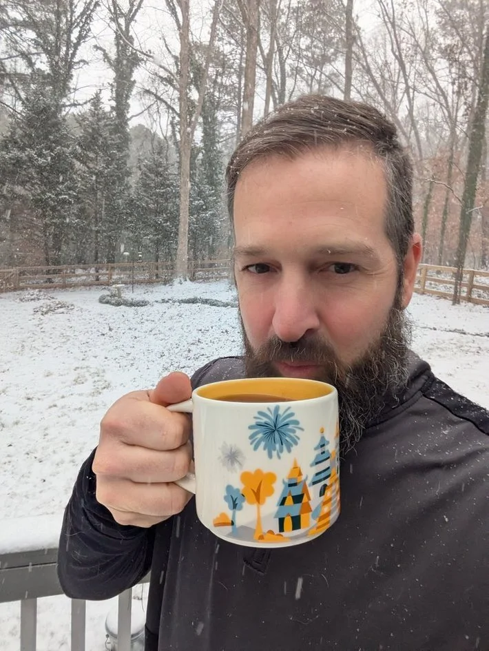 A man with a beard holding a colorful mug with a hot beverage, standing outdoors in a snowy backyard with trees and a wooden fence.