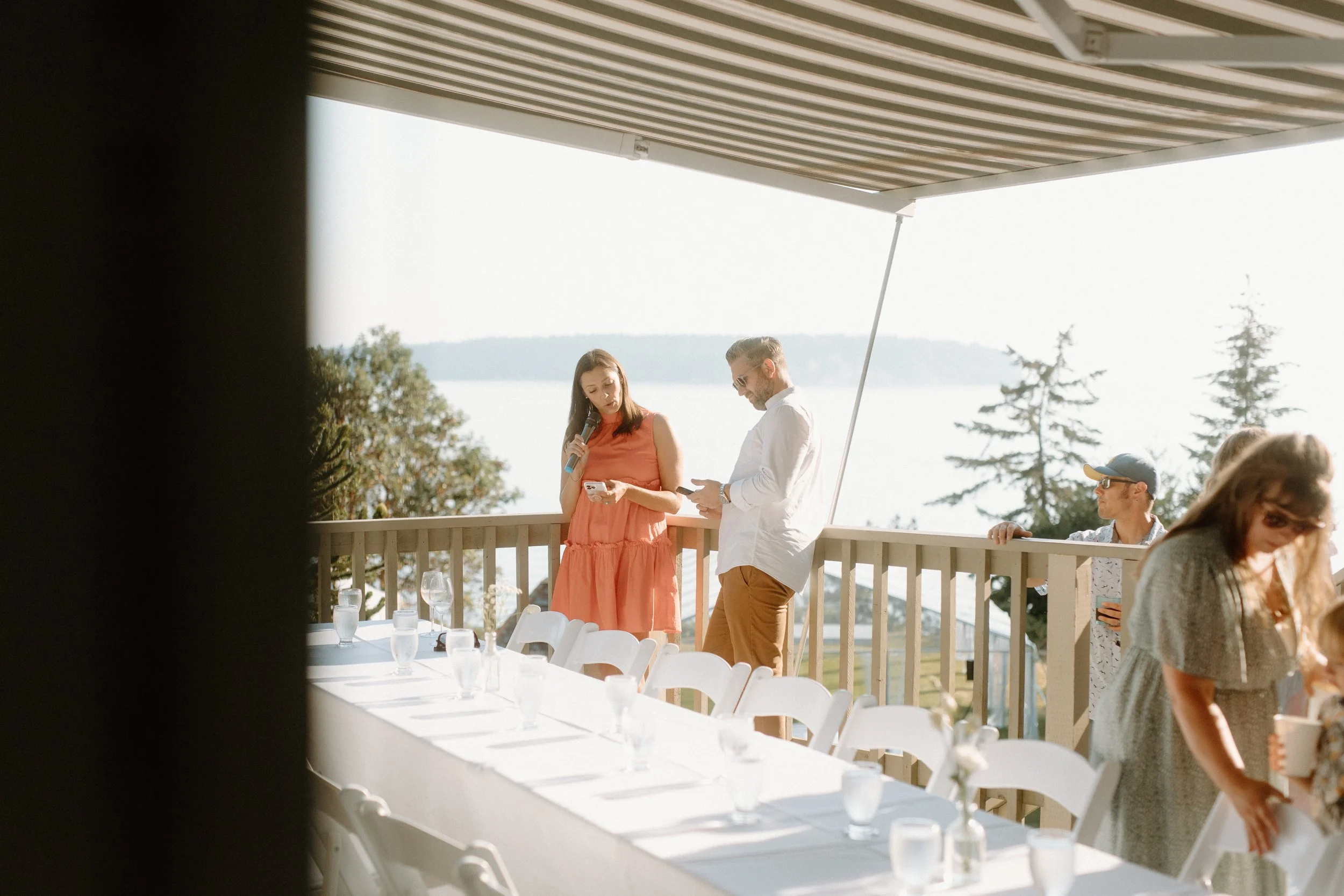 People gathering on a balcony near a table with drinking glasses and water bottles, overlooking a body of water with trees in the background, during a sunny day.