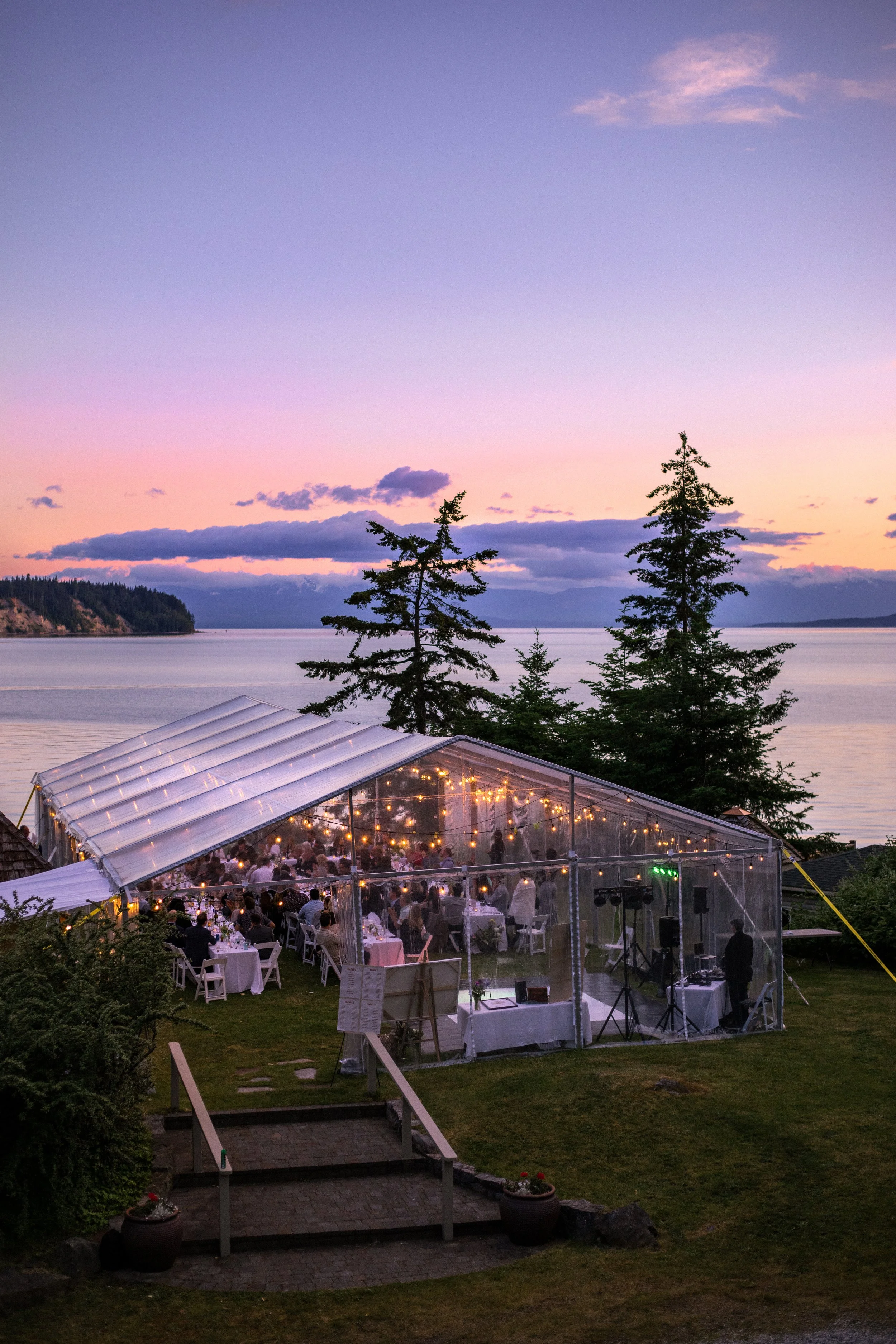 Outdoor wedding reception under a transparent tent with string lights, overlooking a body of water at sunset, with guests seated at tables.