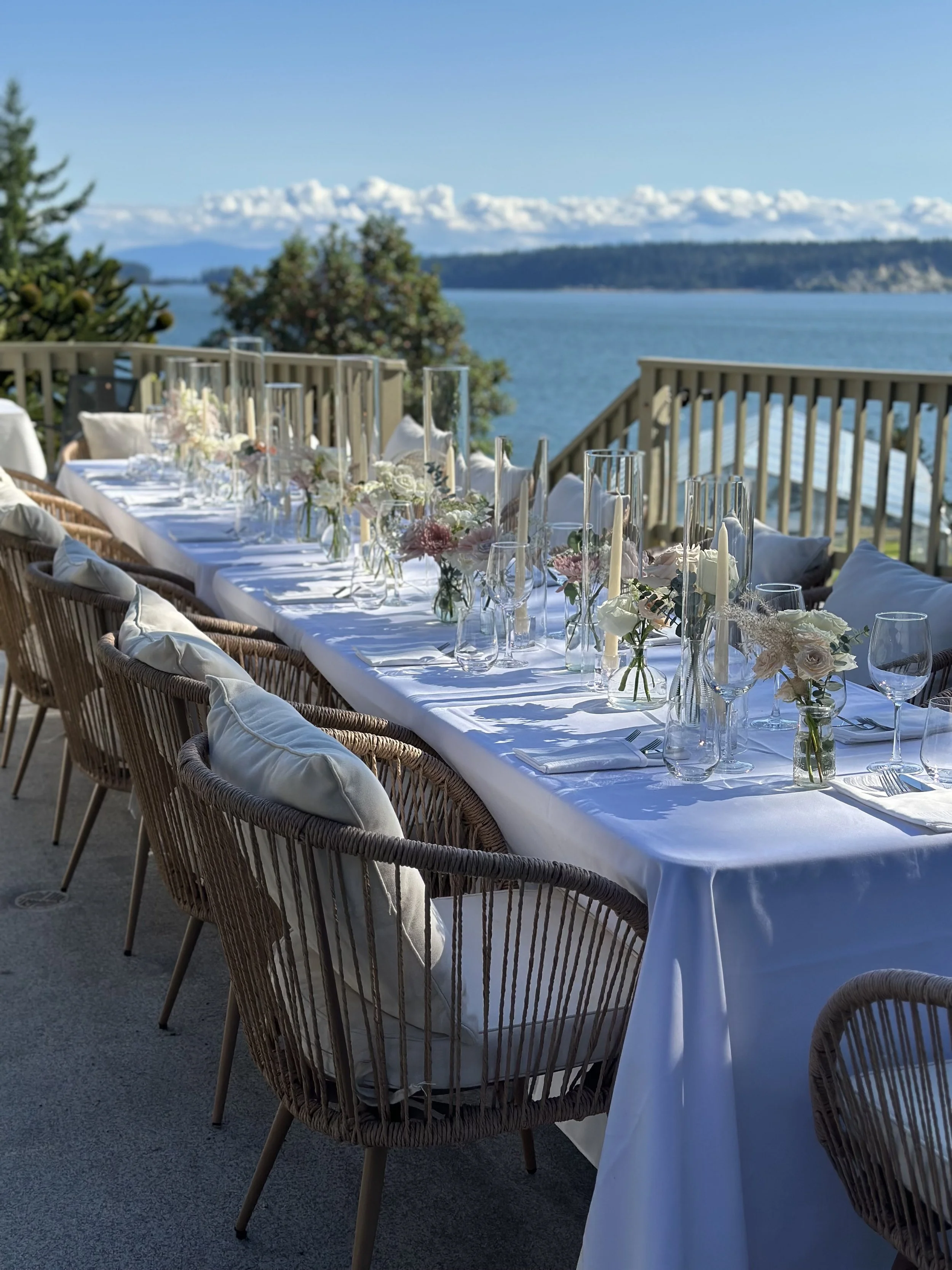 Outdoor dining table set with white tablecloth, glassware, and floral centerpieces, overlooking a body of water with mountains in the background.