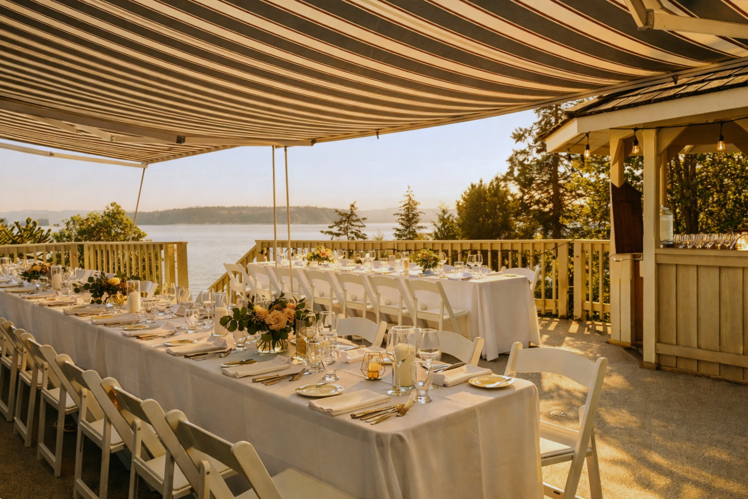 Elegant outdoor dining area set for a celebration with a view of the water, featuring a long table with white tablecloth, floral centerpieces, candles, and arranged place settings under a striped awning.
