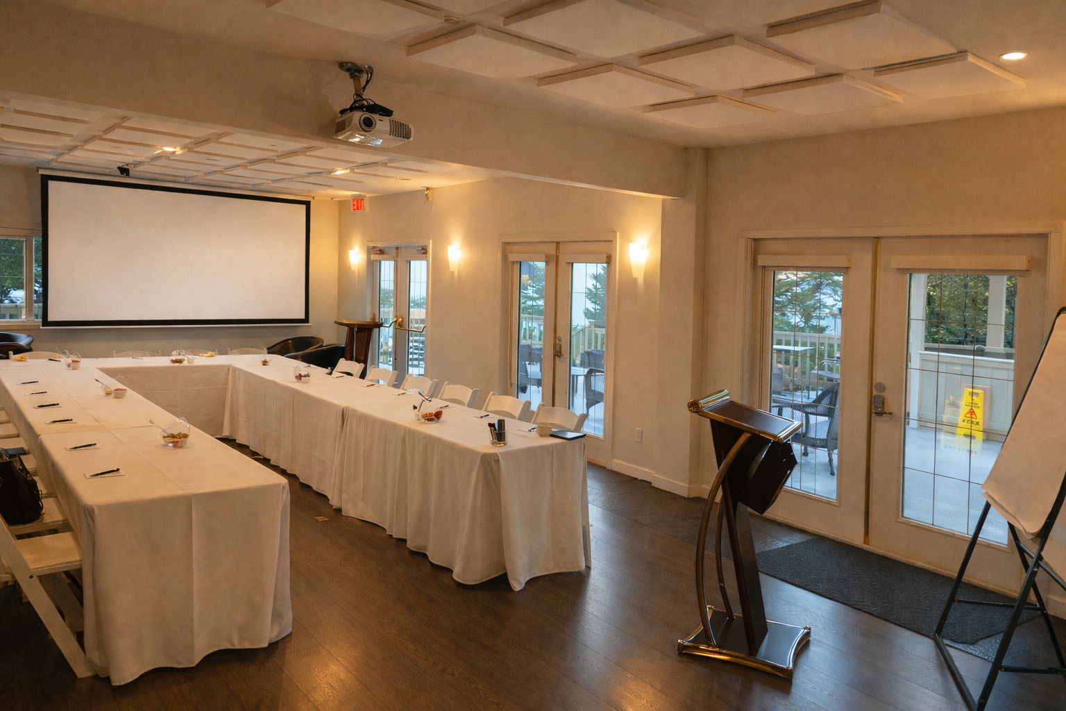Empty conference room set up with a U-shaped arrangement of white-draped tables, a large projector screen, a podium, and a flip chart, with some boxes of snacks on the tables and chairs around the room, outdoor seating visible through the windows and doors.