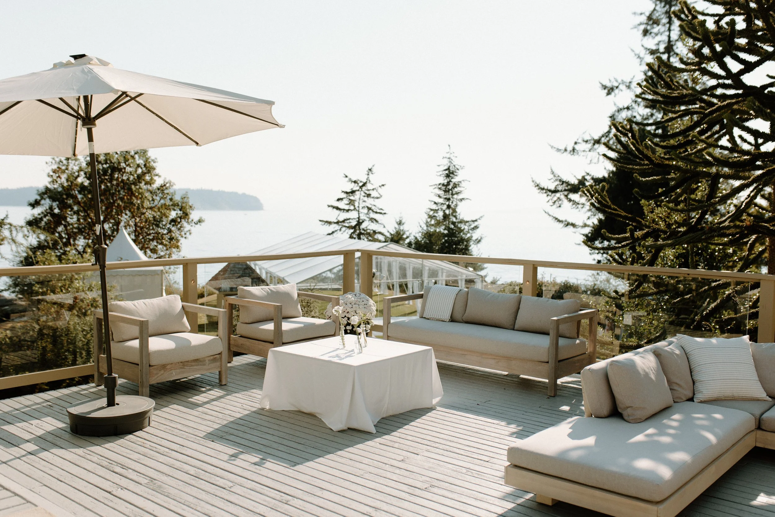 Outdoor patio with beige seating, white table with flowers, large umbrella, overlooking trees and water in the distance on a sunny day.