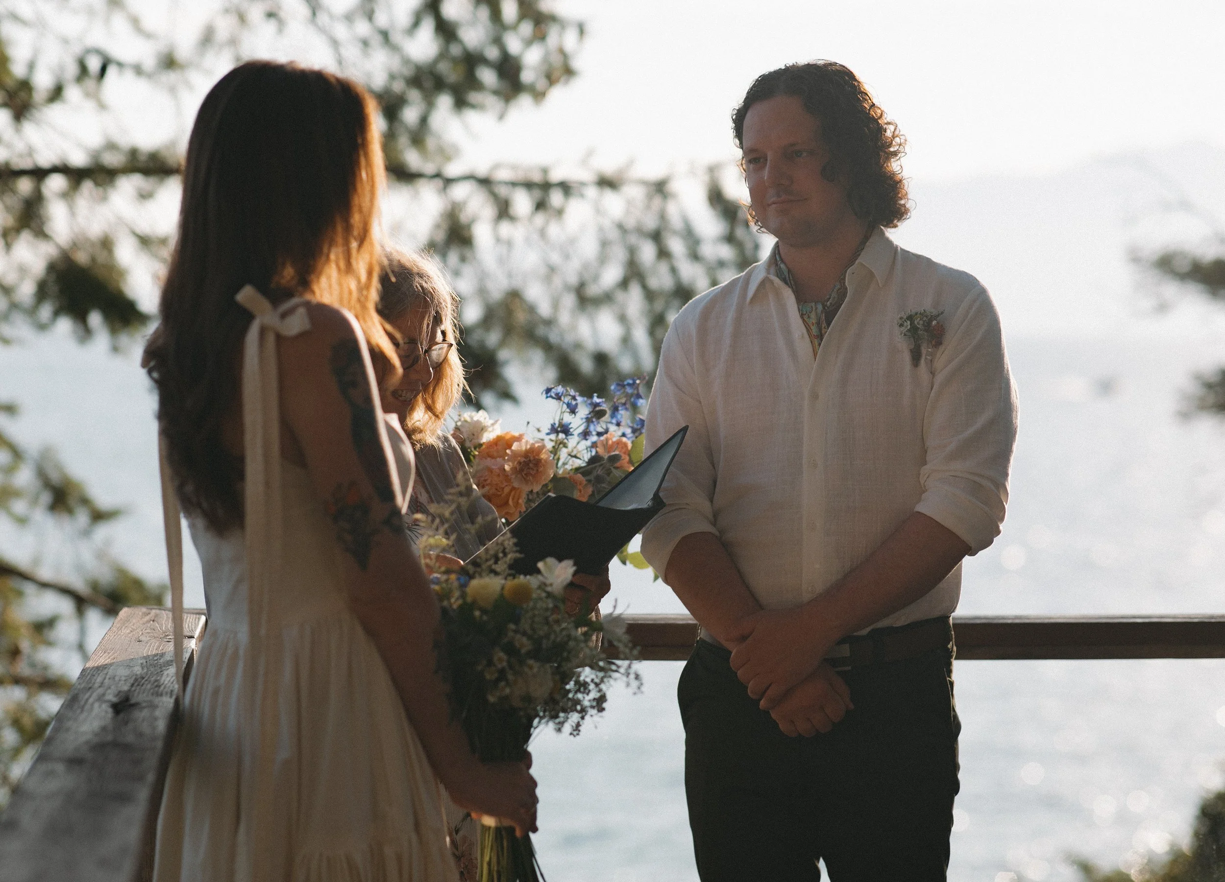 A couple is standing outdoors during a ceremony, with a woman officiating and holding a certificate or book. The woman on the left has long hair with tattoos on her arms and is holding a bouquet of flowers. The man on the right is wearing a white shirt with rolled-up sleeves and is looking at the woman officiating. The background features trees and bright sunlight.