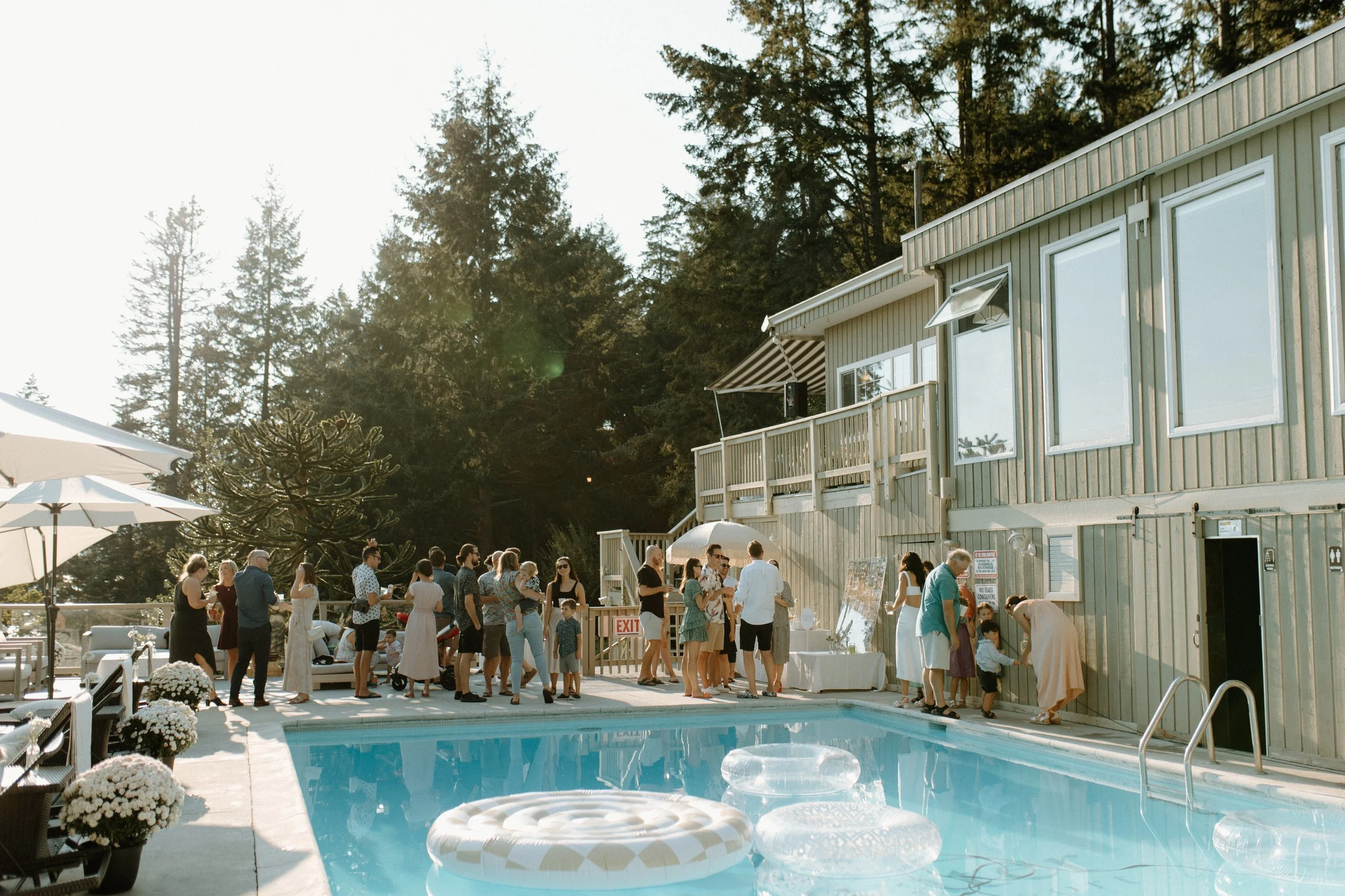 People gathering by a swimming pool at a summer outdoor party, with a wooden building, trees in the background, and sunlight.