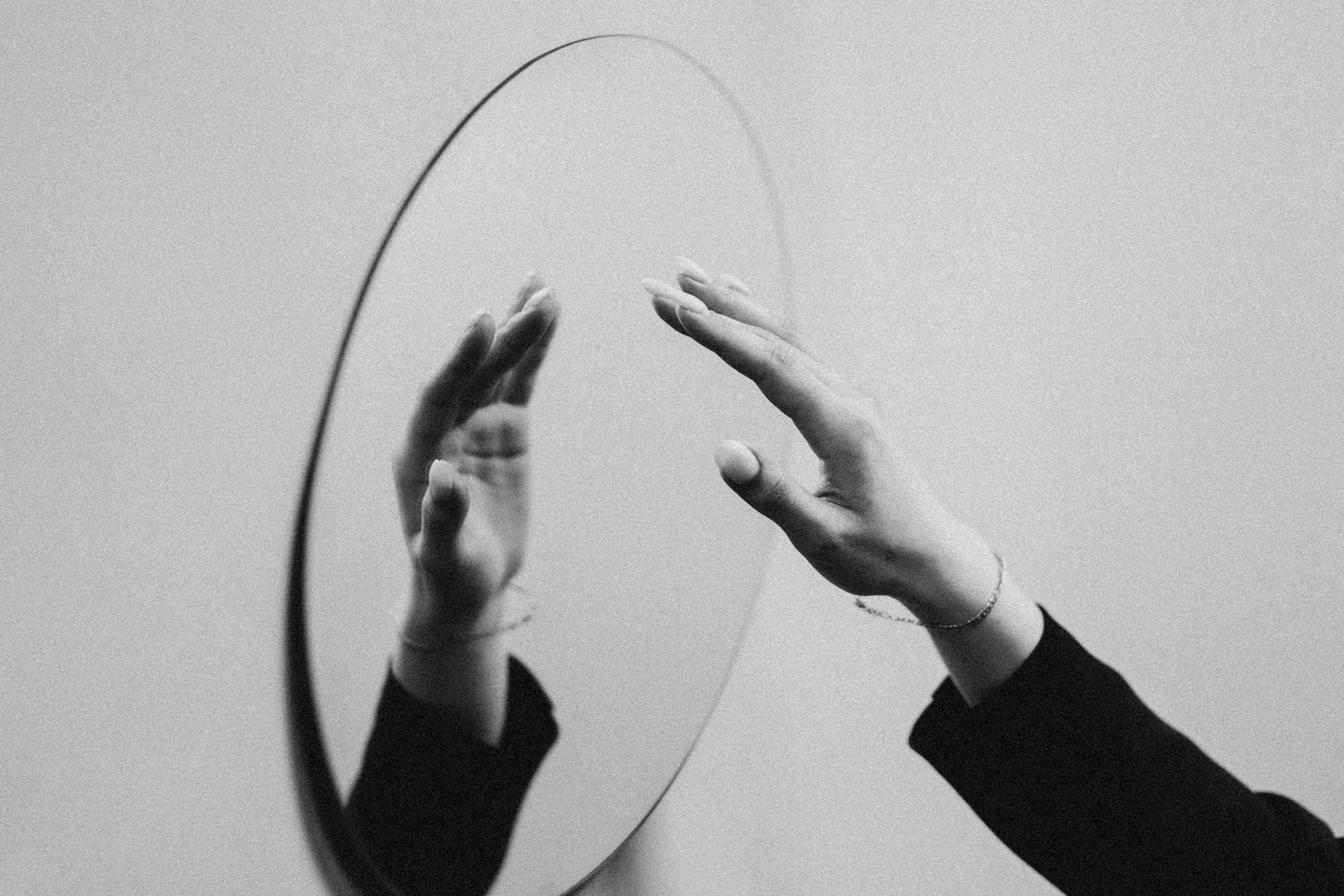 A person's hands reaching towards a round mirror, reflected in the mirror are their fingers and part of their face, black and white photograph.