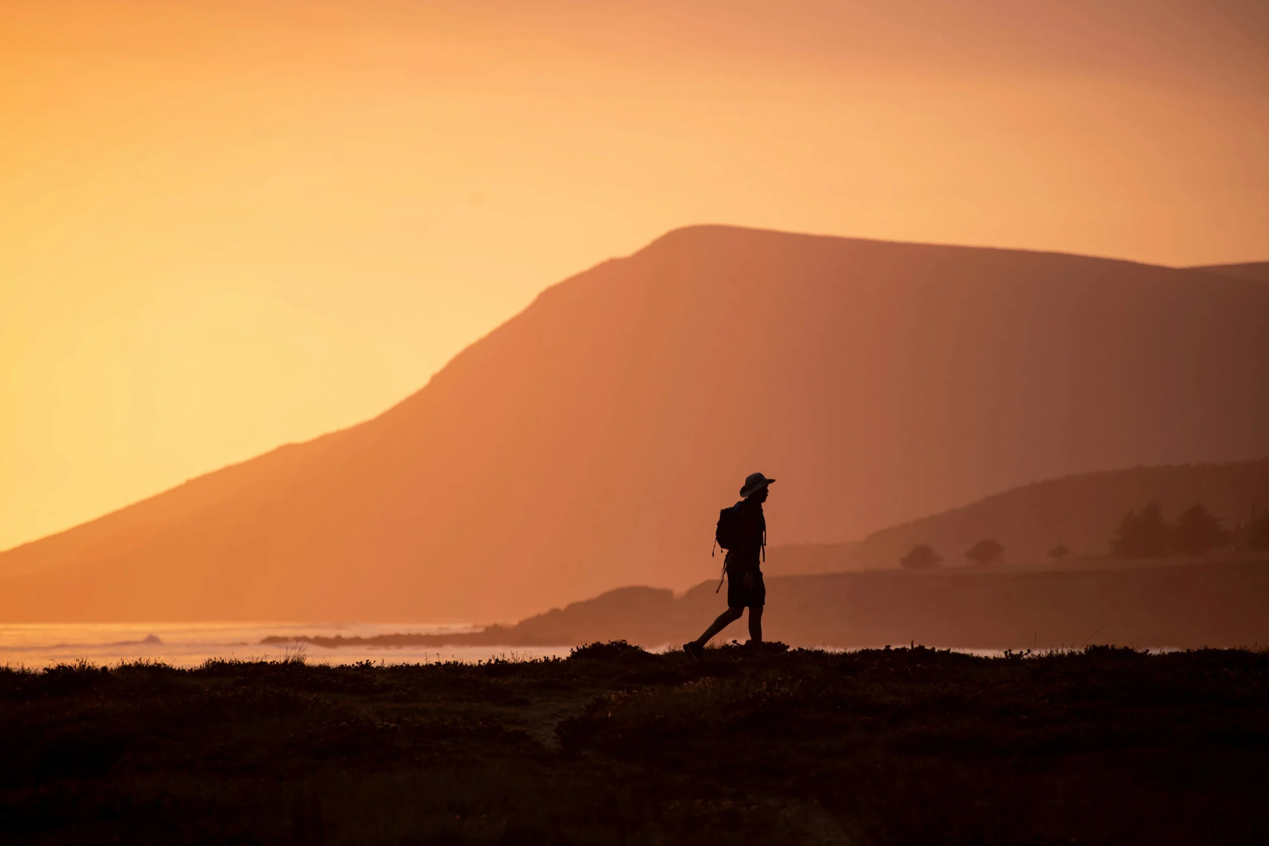 A person hiking along a coastal area during sunset with mountains in the background.