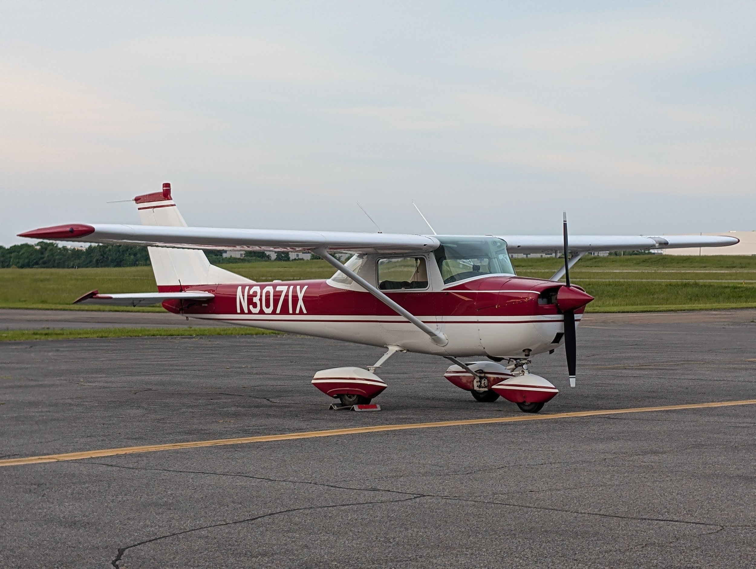 Small red and white single-engine airplane parked on an airport tarmac with grassy fields and a cloudy sky in the background.