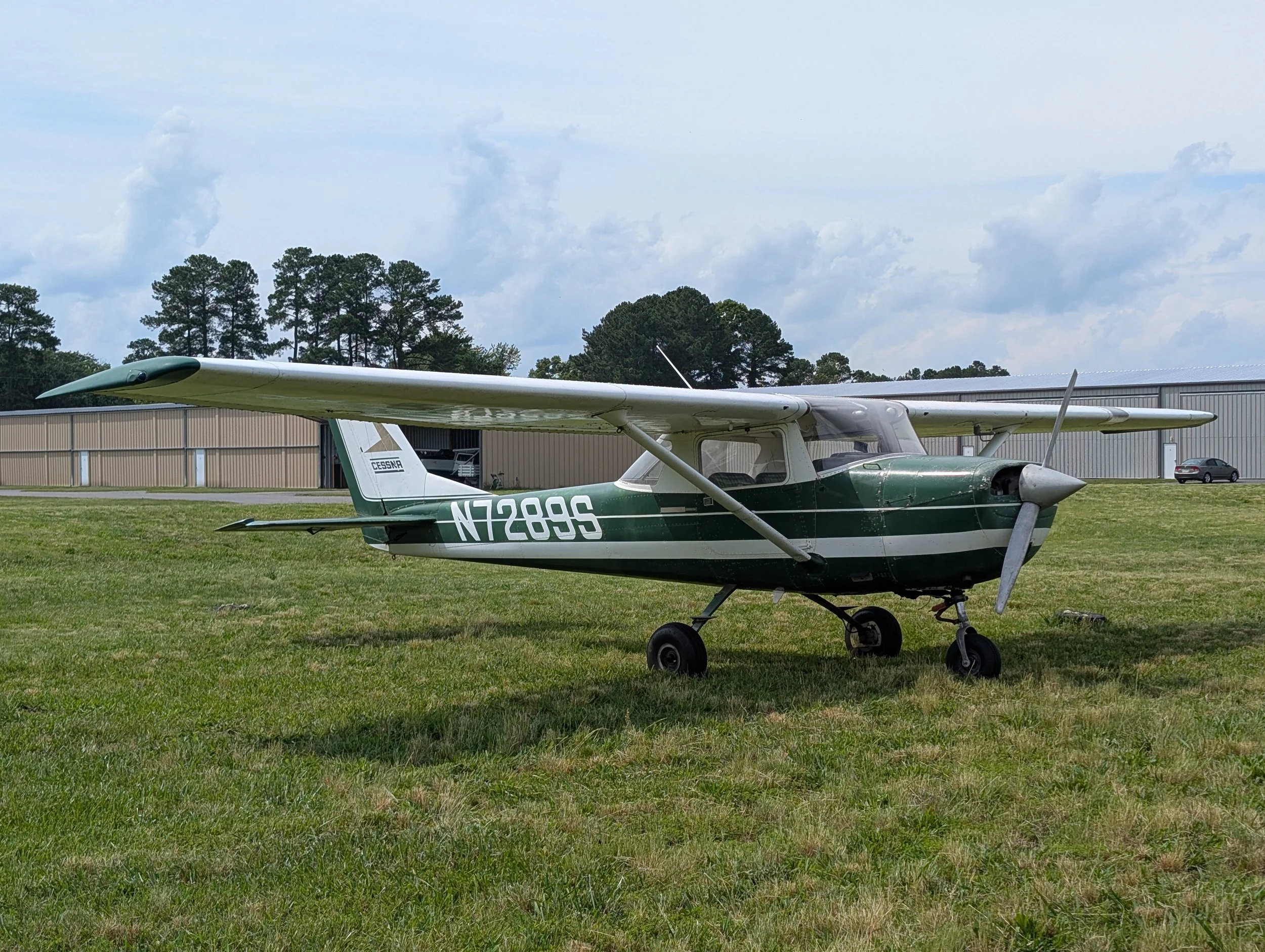 Small green and white Cessna aircraft parked on grassy field near hangar with cars in background.