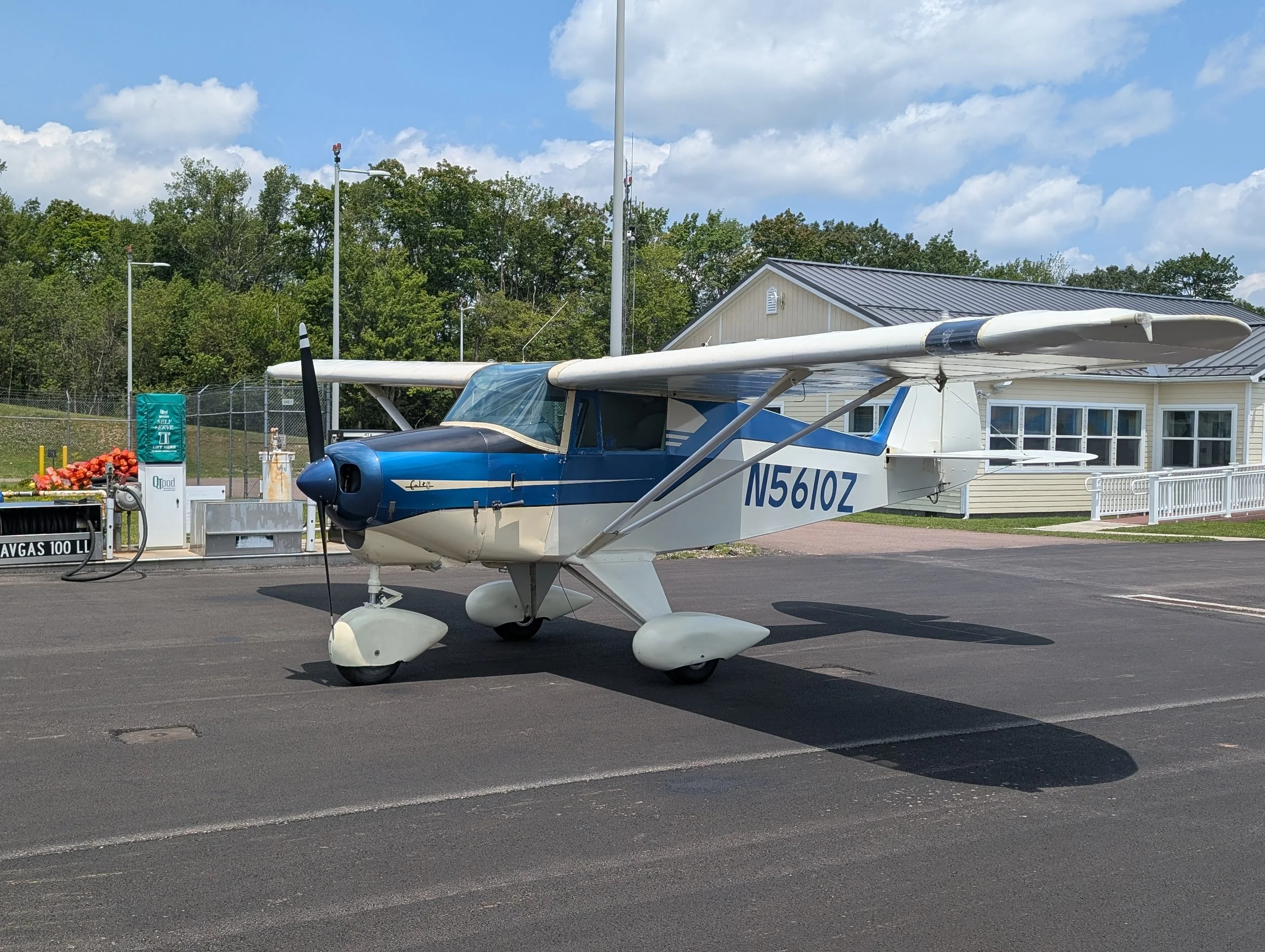 Small white and blue single-engine aircraft parked on tarmac near fuel pumps at an airport.