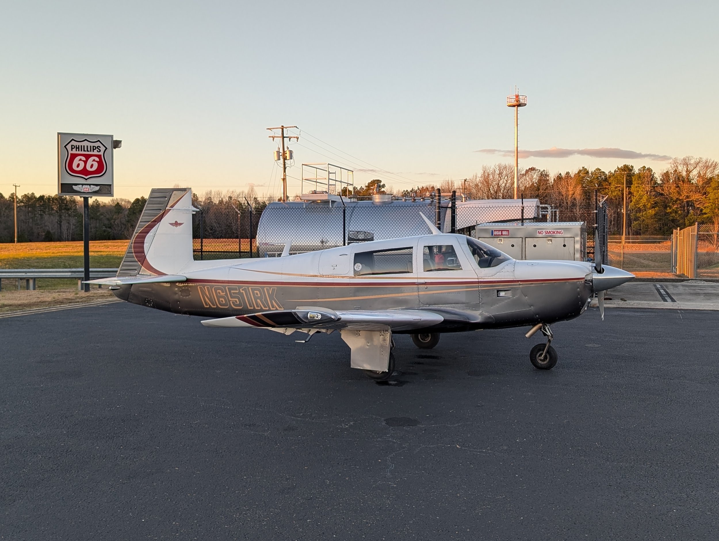 Small aircraft parked on a tarmac near a Phillips 66 gas station sign, with a background of trees and utility poles during sunset.