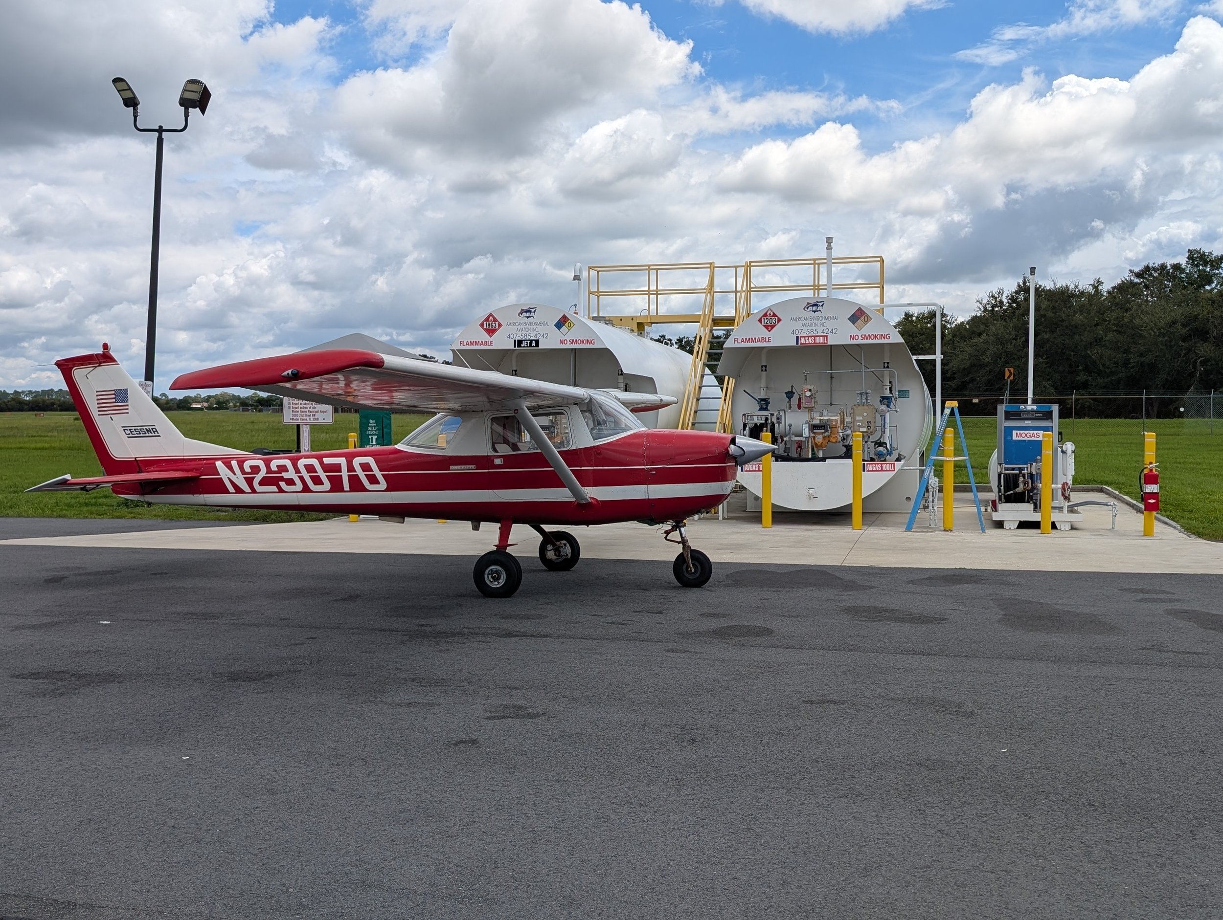 Small red and white aircraft parked in front of fuel tanks at an airport, with a cloudy sky overhead.