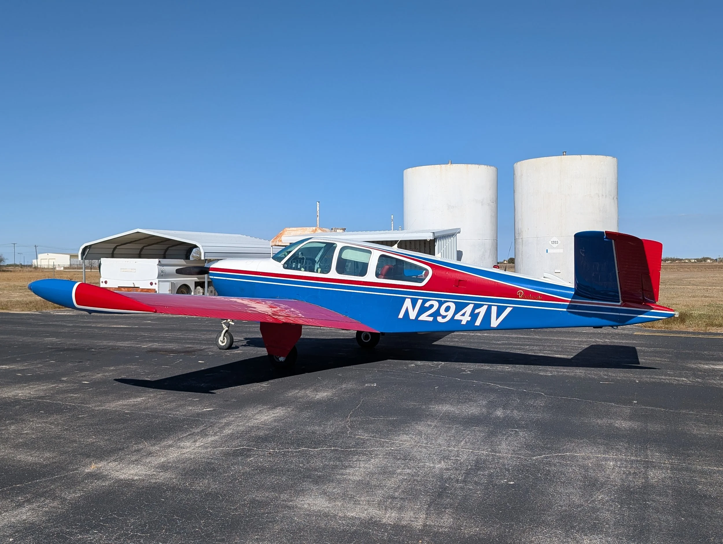 A small blue, red, and white aircraft parked on an asphalt tarmac, with two large white storage tanks and a hangar in the background.