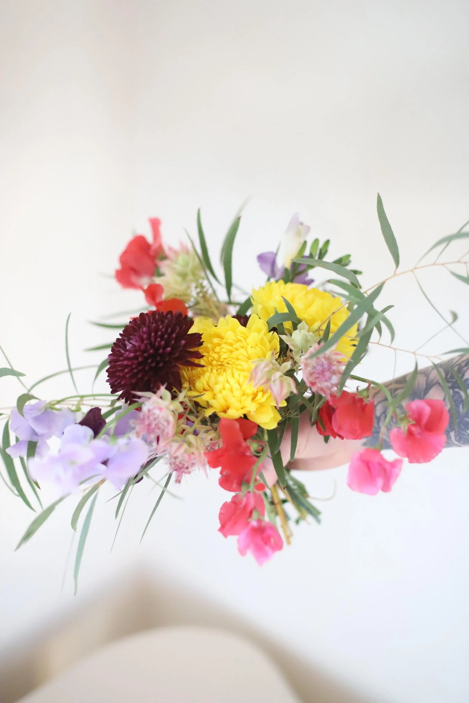 Colorful bouquet of flowers with yellow, purple, red, pink, and maroon blossoms and green foliage on a white background.