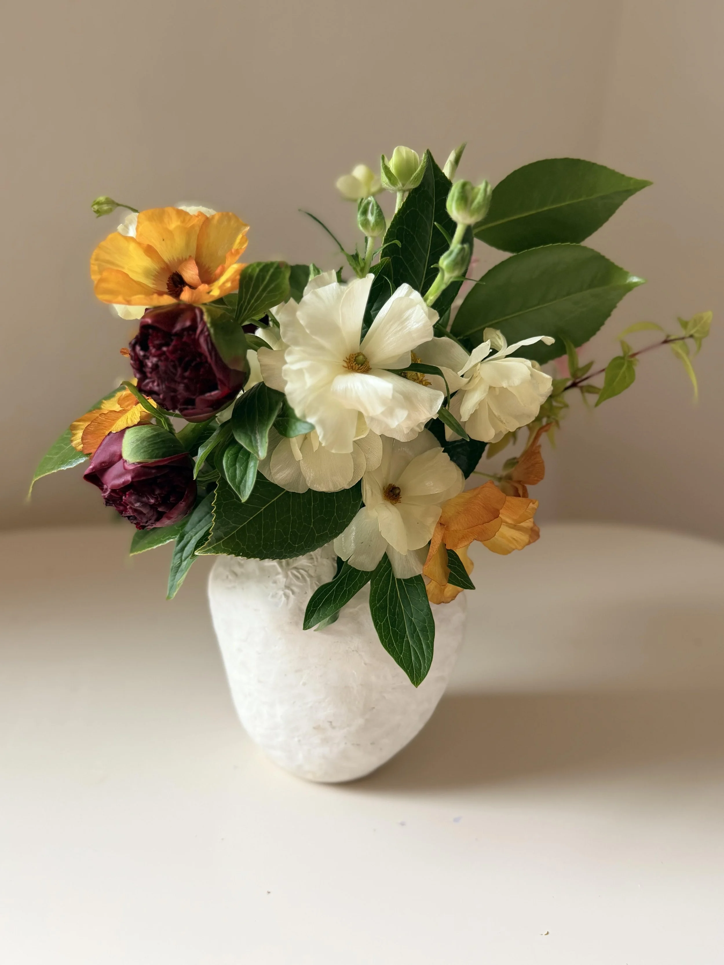 A vase with white, yellow, and dark red flowers and green leaves on a beige surface.