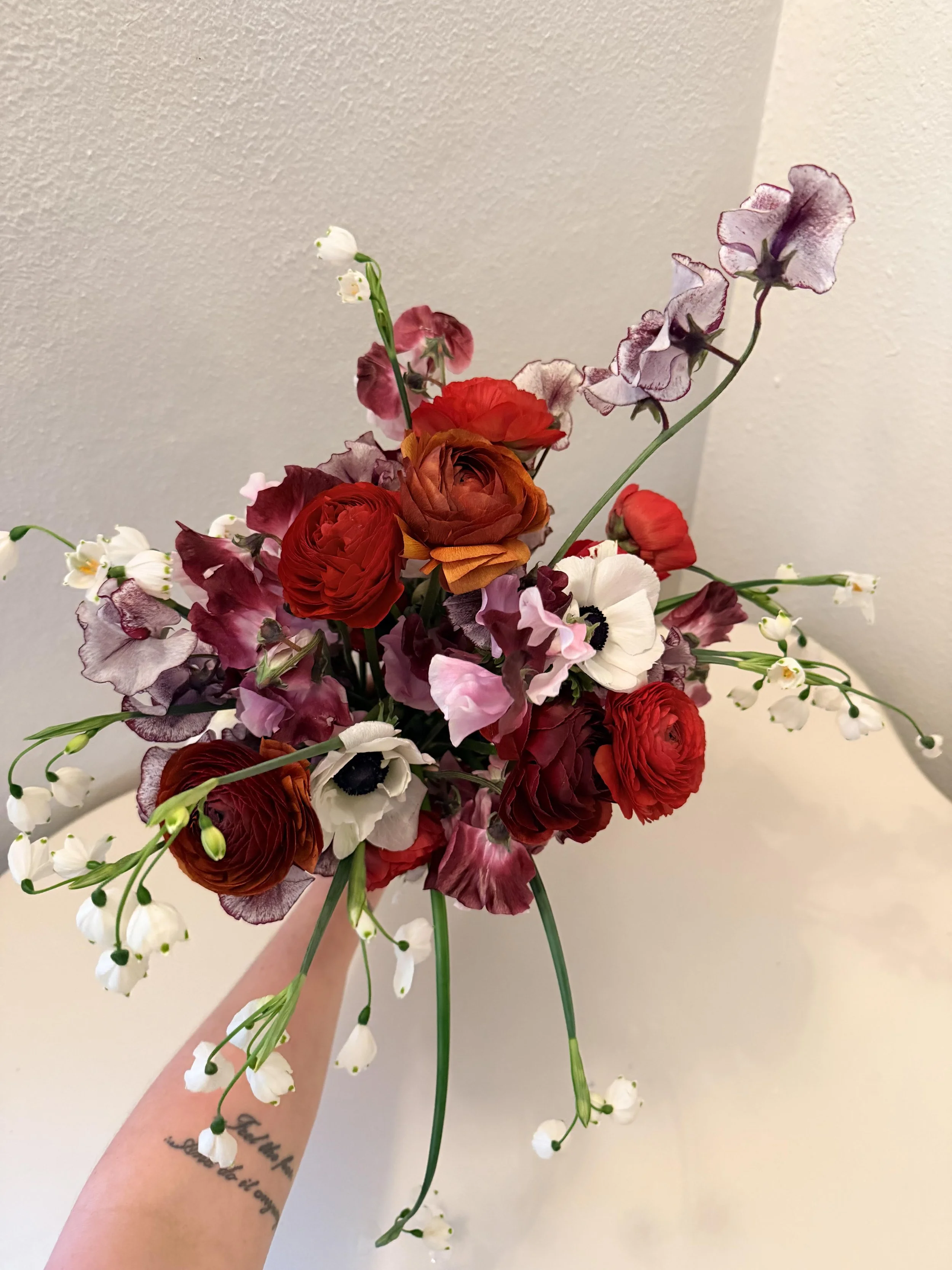 A hand holding a colorful bouquet of various flowers including white, red, pink, purple, and orange blooms against a plain wall background.