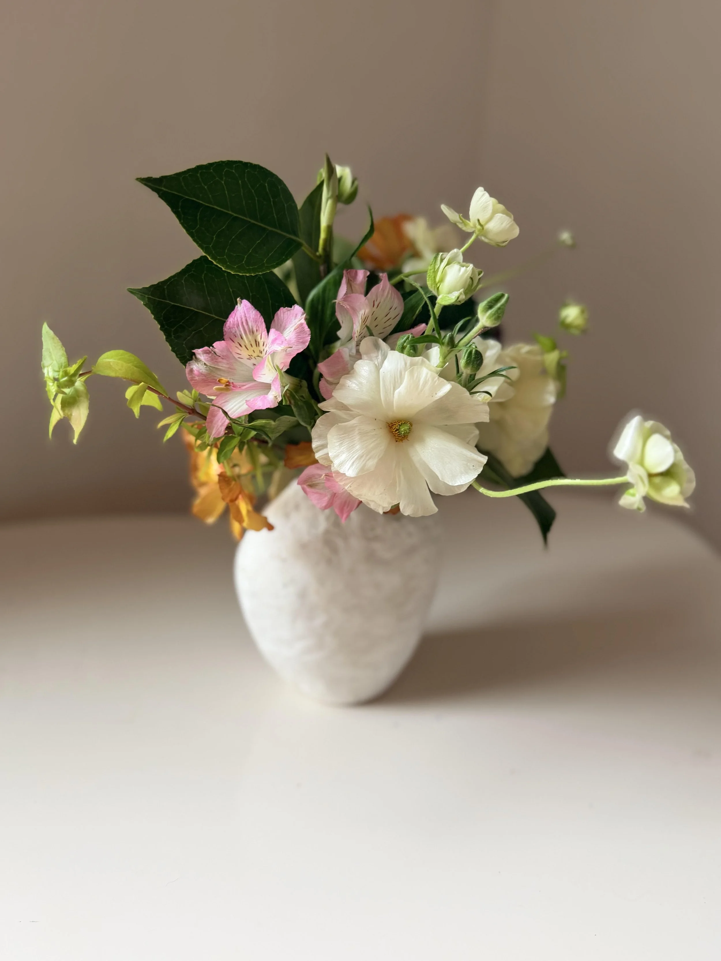 A white ceramic vase holding a bouquet of flowers including white, pink, yellow, and orange blossoms with green leaves.