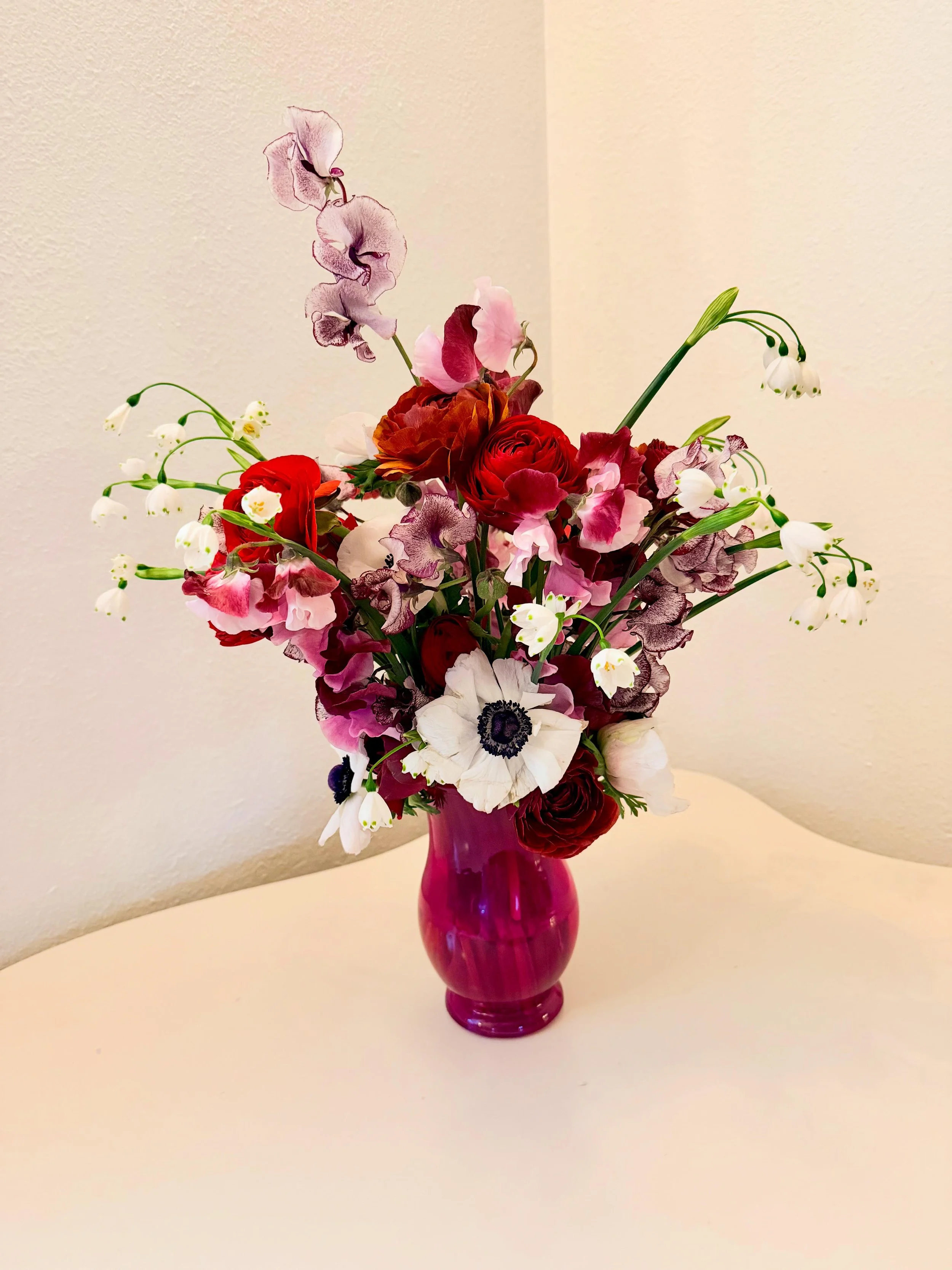 A bouquet of various flowers in a pink vase on a white surface, with a plain white wall background.