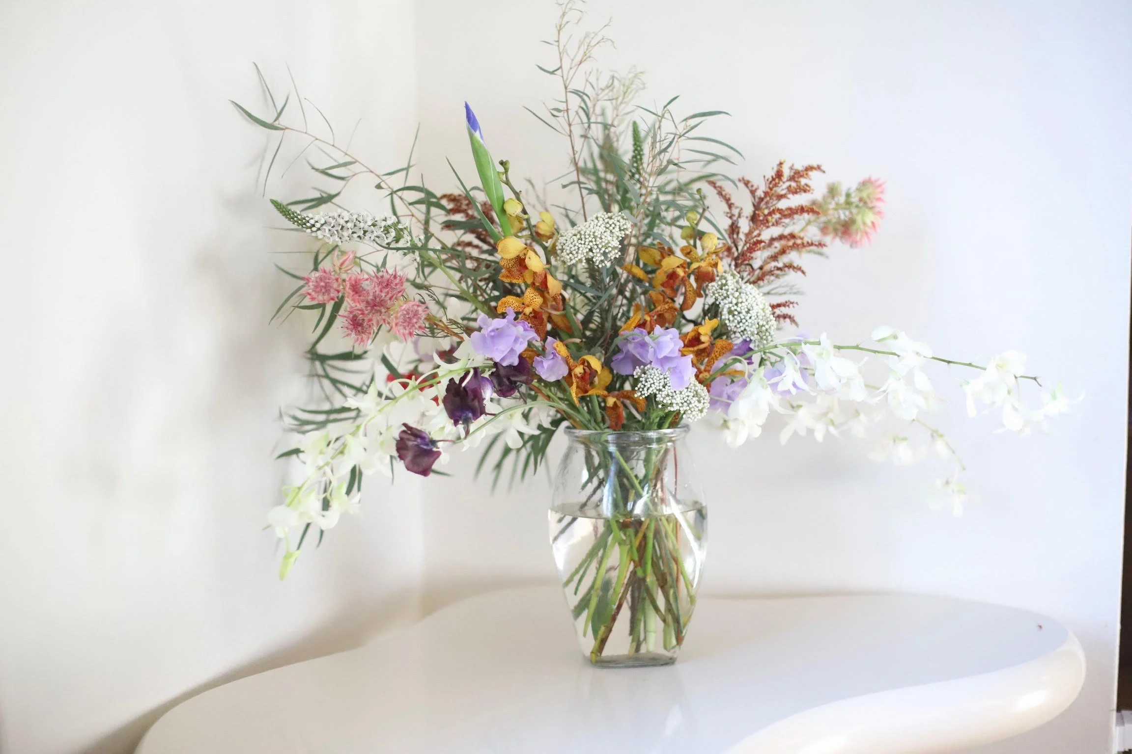 A colorful bouquet of various flowers in a clear glass vase on a white table against a plain white wall.
