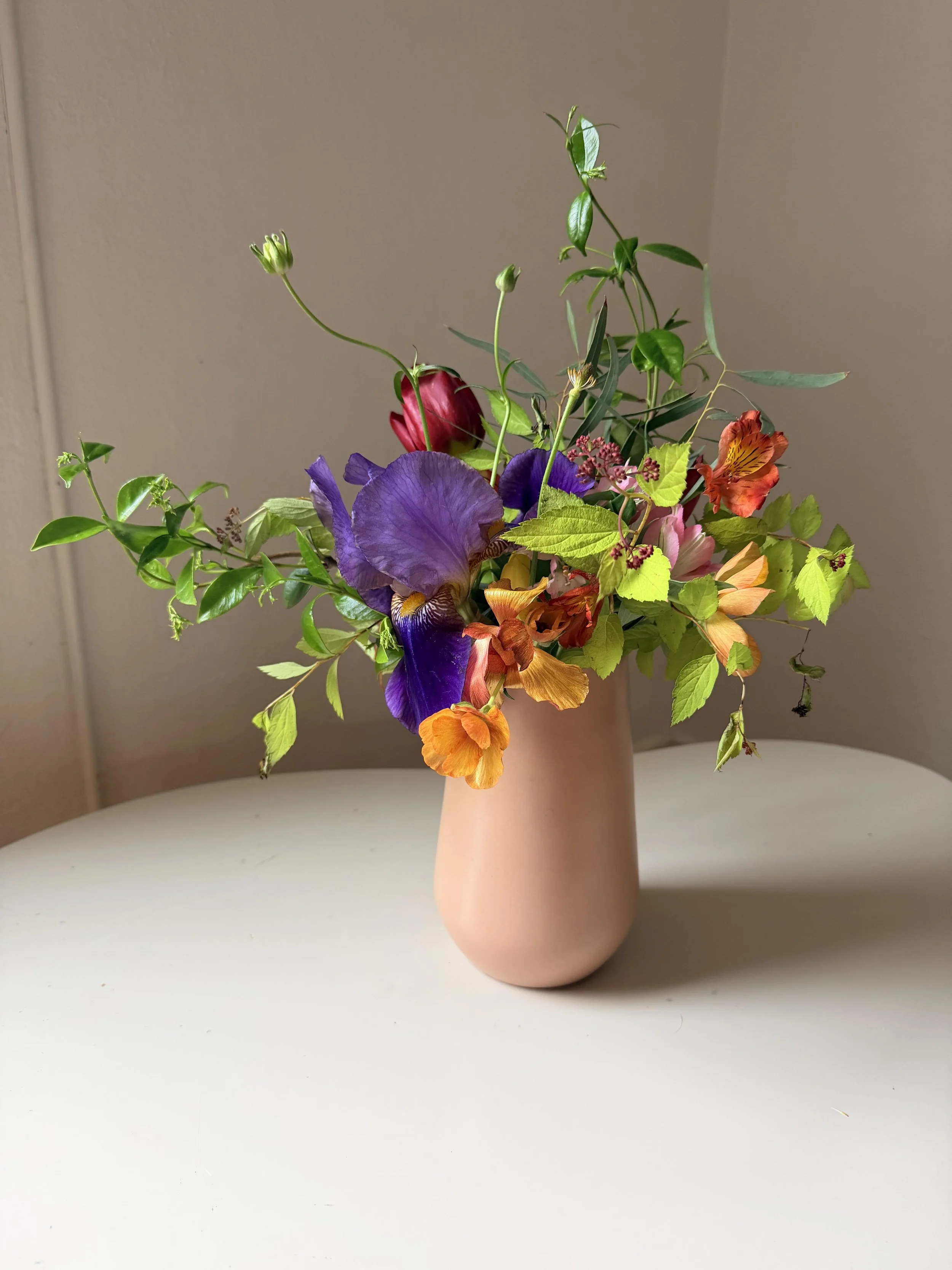 A pink ceramic vase filled with a colorful assortment of flowers and greenery, sitting on a light-colored table against a plain wall background.