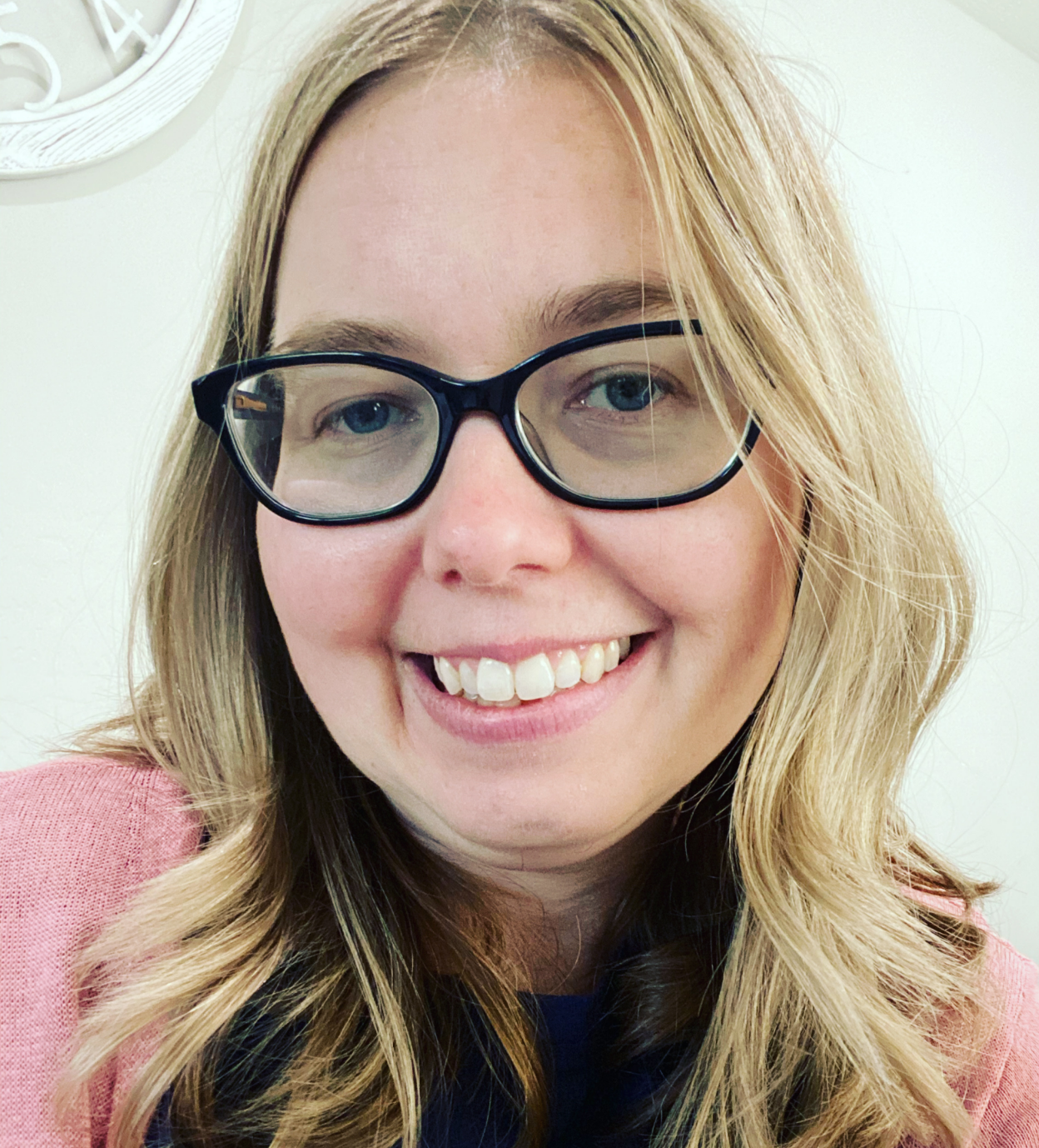 Close-up photo of a woman with blonde wavy hair, wearing large black glasses, smiling showing teeth, and dressed in a pink blazer and navy top.