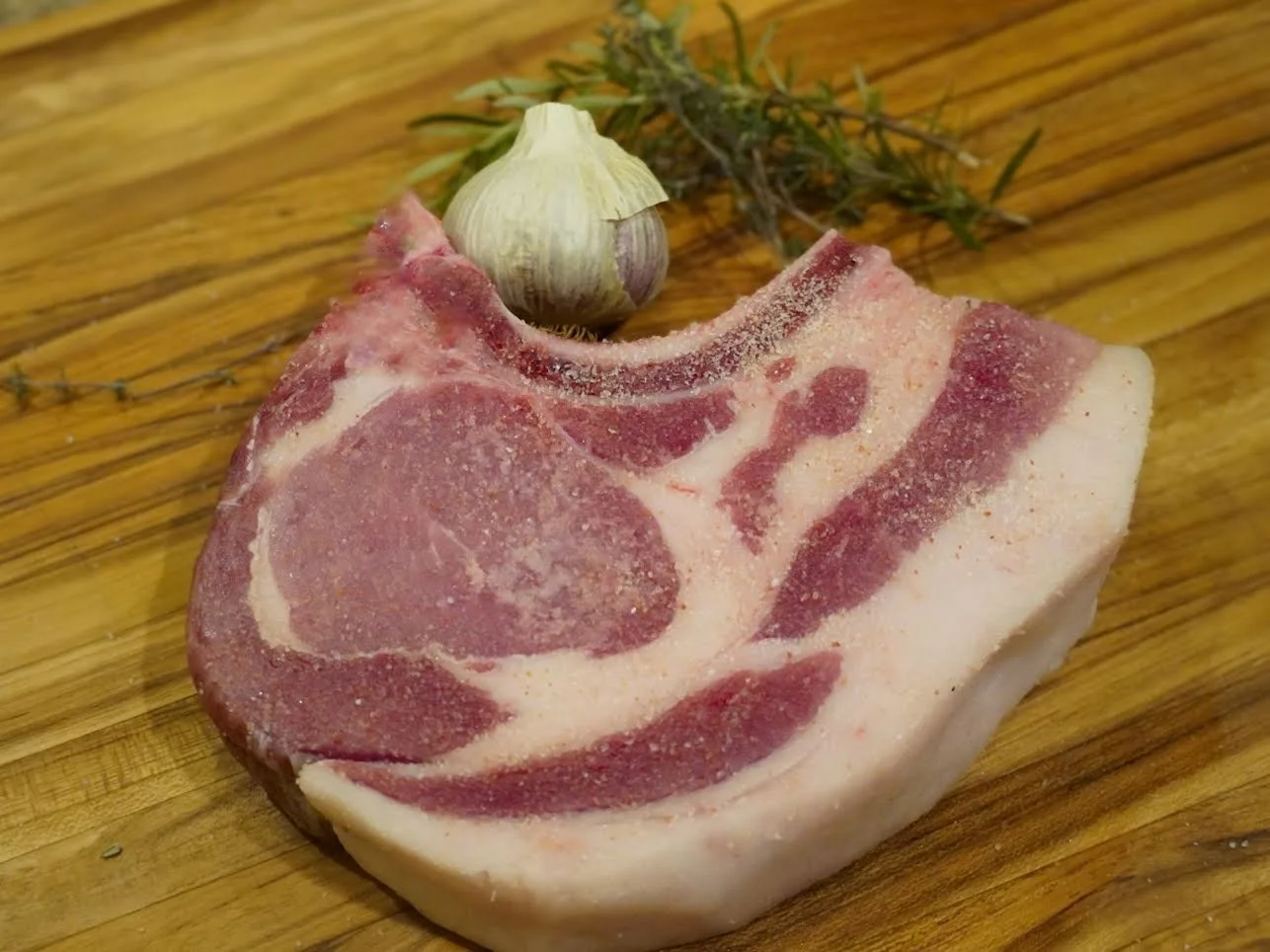 Raw bone-in pork chop on wooden surface with garlic bulb and sprigs of herbs.