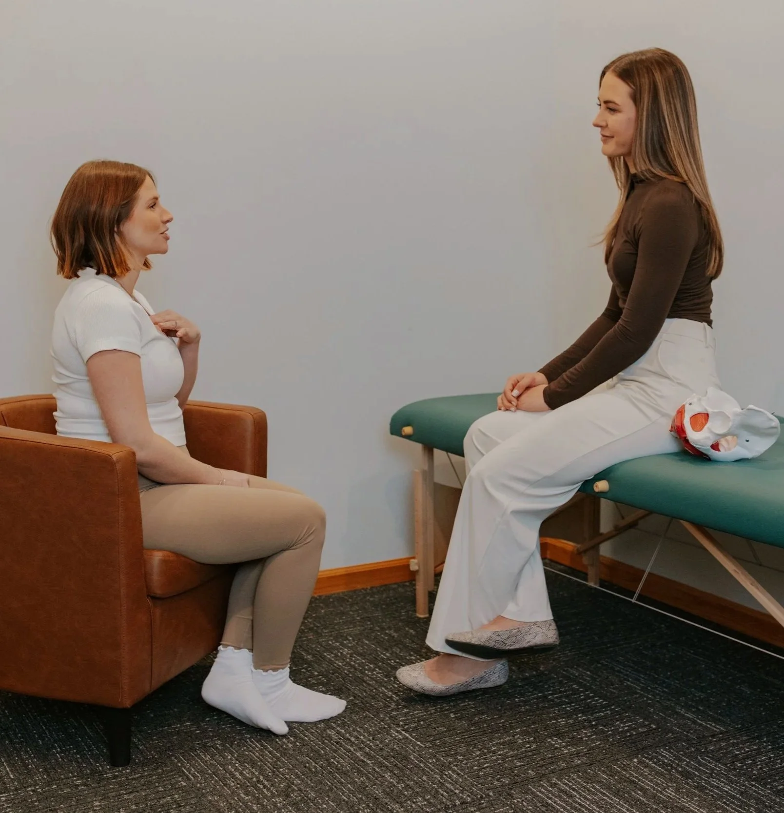 Two women engaged in a consultation, one sitting on a brown armchair and the other on an examination table, in a clinical setting.
