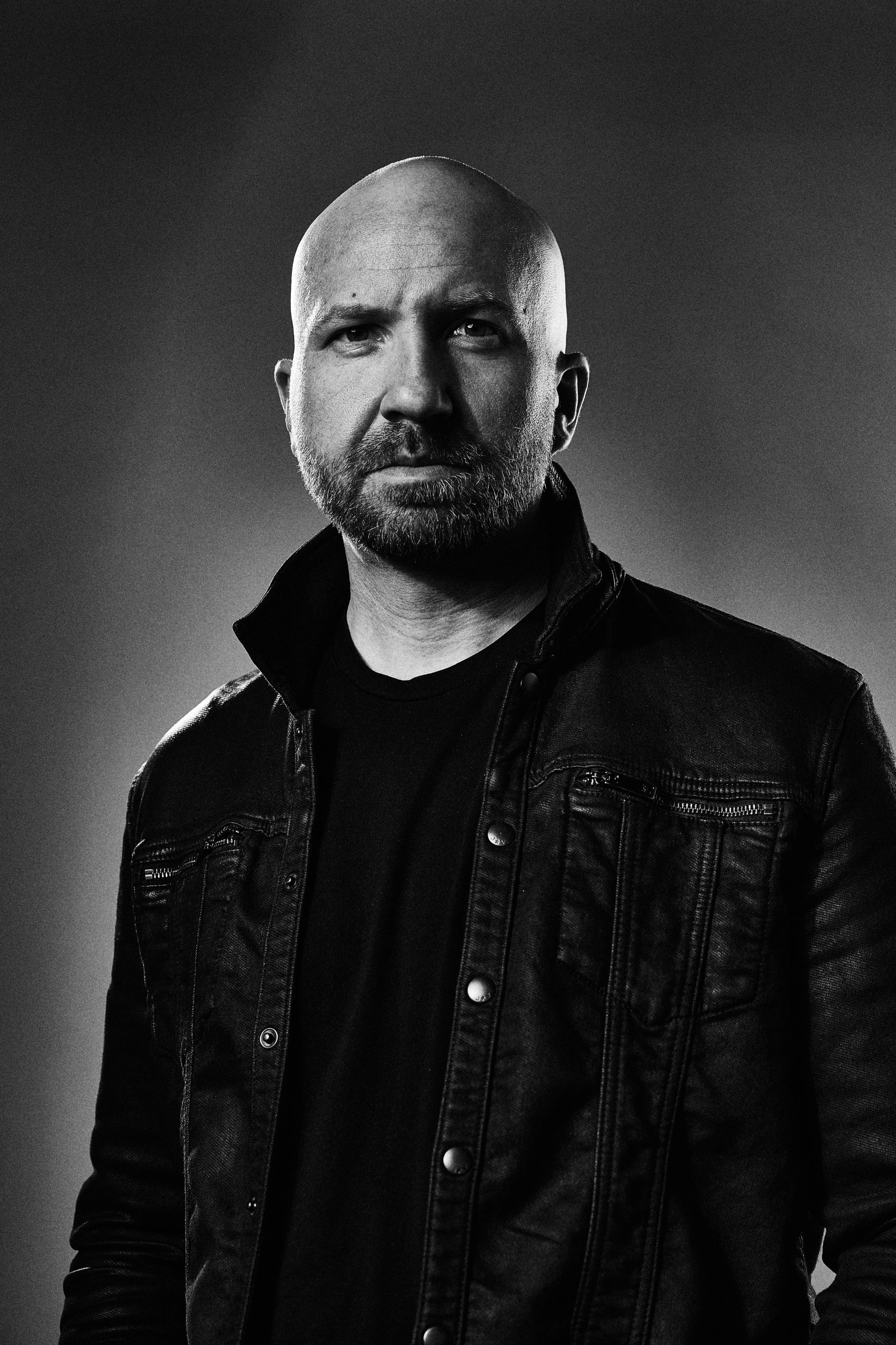 Black and white photo of a bald man with a beard wearing a black leather jacket and shirt, looking directly at the camera.