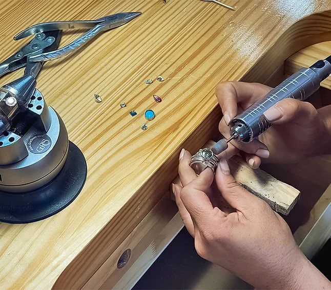 A person jewelry crafting, holding a small tool and working on a ring at a jewelry workbench. The workbench also has assorted small gemstones and jewelry-making tools.