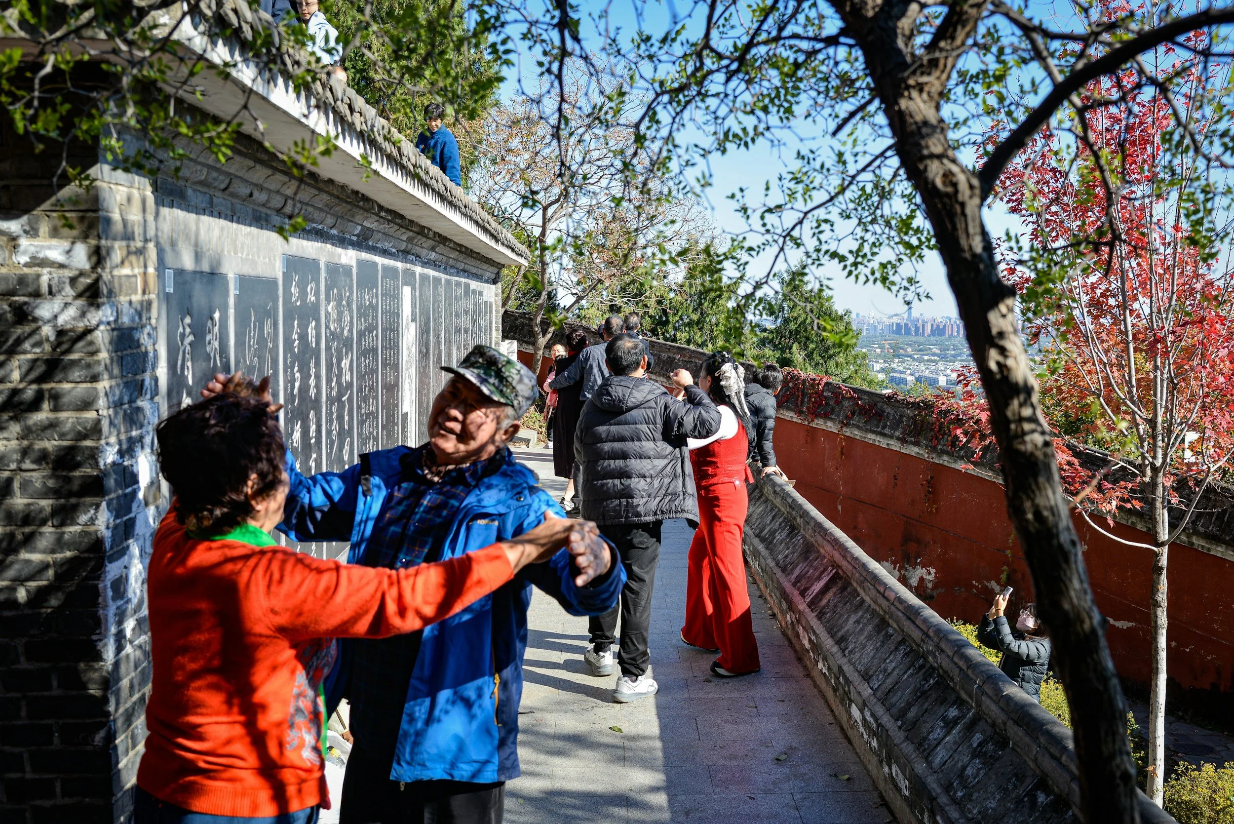 People standing on a bridge, some taking photos and looking at a wall with inscriptions, with trees and a cityscape in the background on a sunny day.