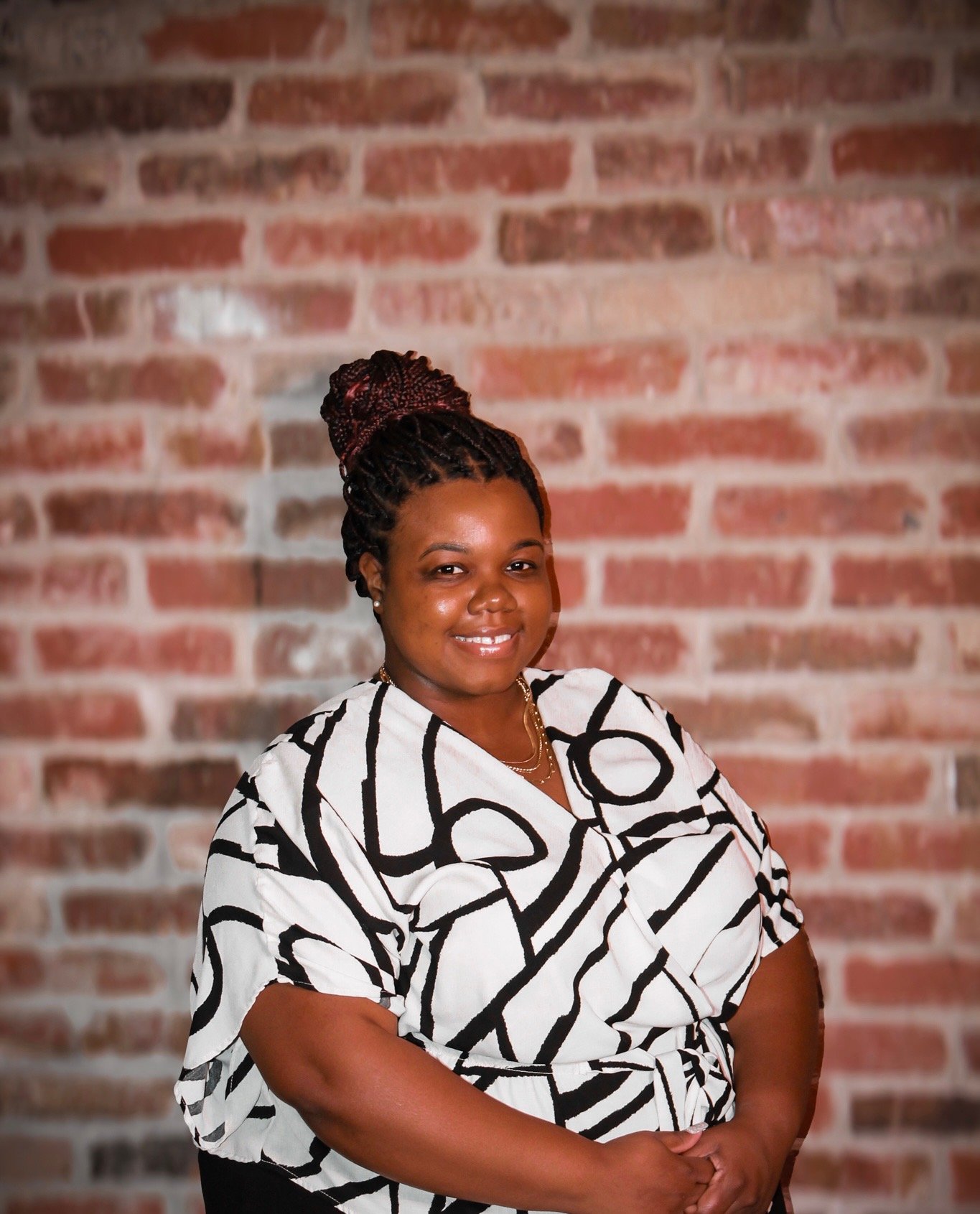 A woman with braided hair styled in a bun wearing a black and white patterned shirt, standing in front of a brick wall with a friendly smile.