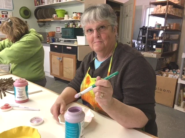 Older woman with gray hair and glasses sitting at a table, holding a paintbrush, in a craft or art studio. She wears an apron and looks at the camera. Shelves and another person in a green hoodie are visible in the background.