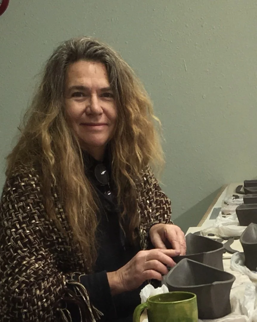 A woman with long, wavy brown hair sitting at a table, working on pottery molds with various pottery pieces and tools around her.