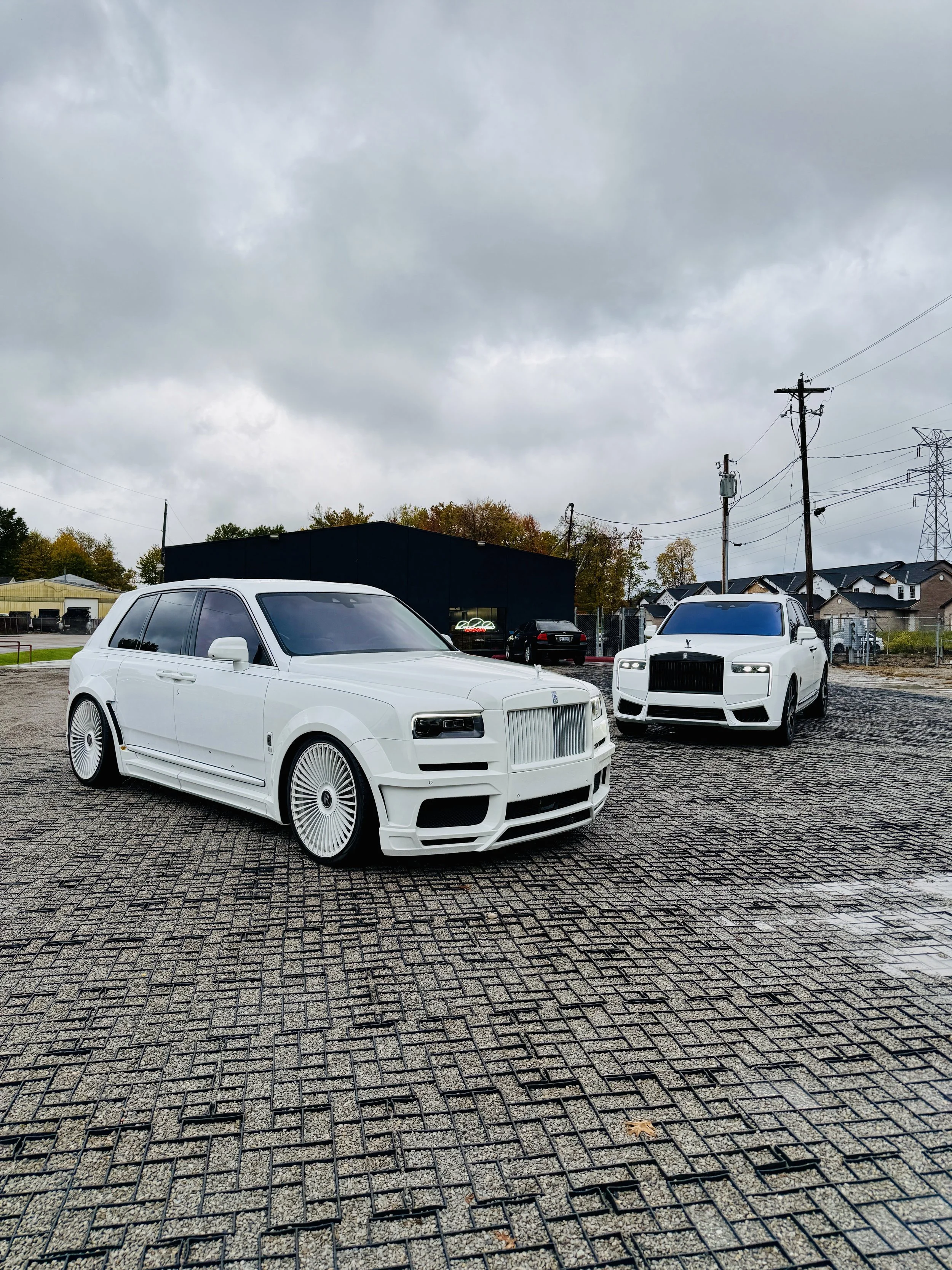 Two white luxury cars parked on a brick pavement outside on a cloudy day.