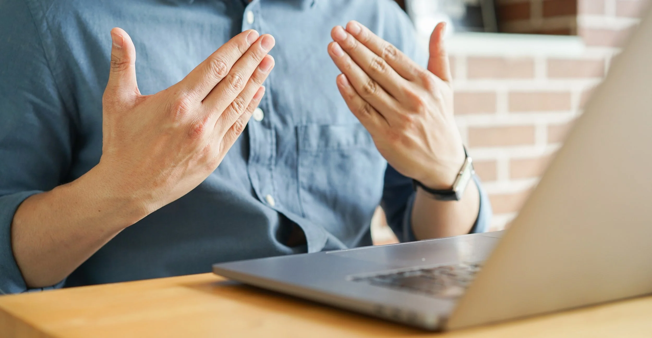 Person gesturing with hands while sitting at a table with a laptop, brick wall background.