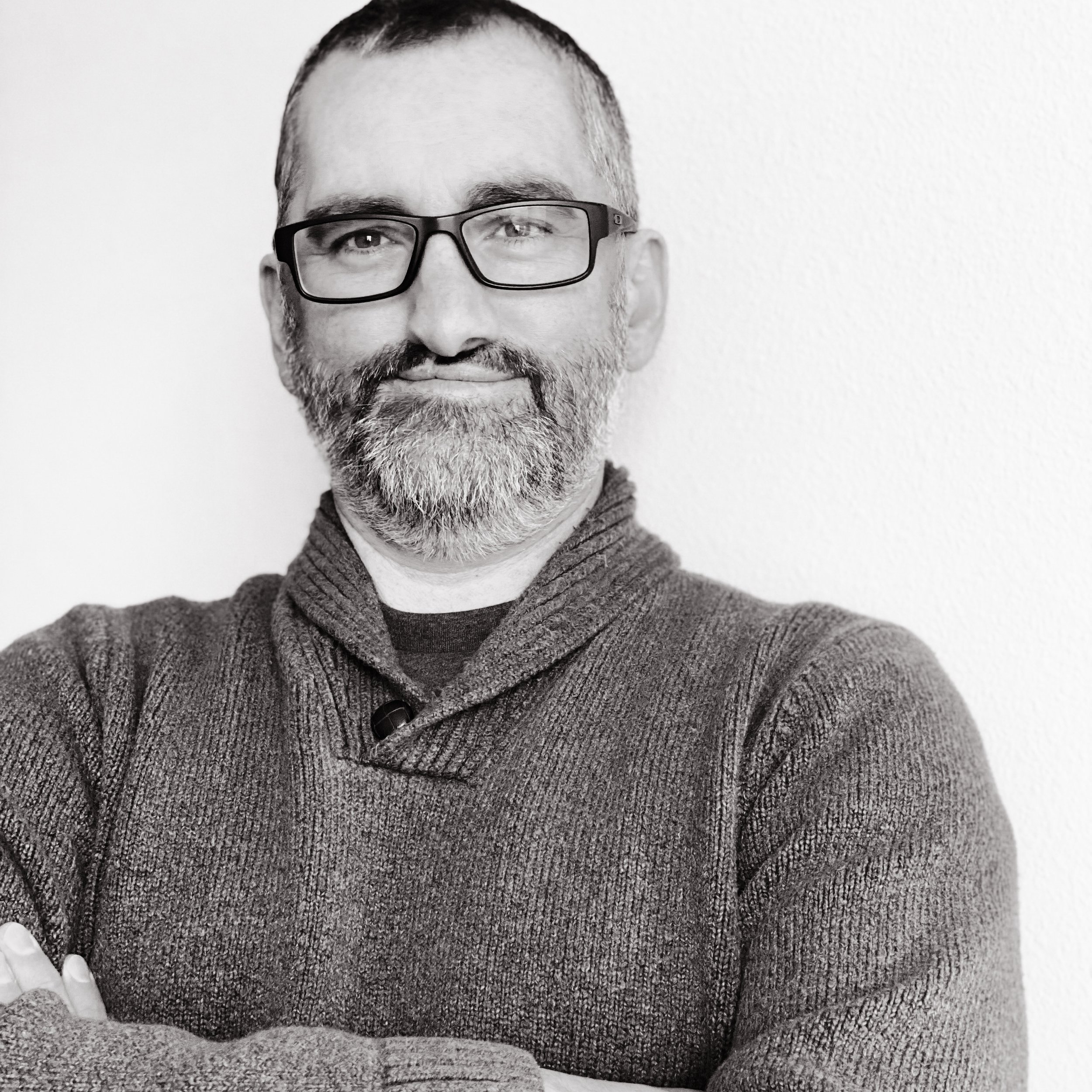Black and white photo of a bearded man with glasses, smiling, arms crossed, wearing a sweater, against a plain background.